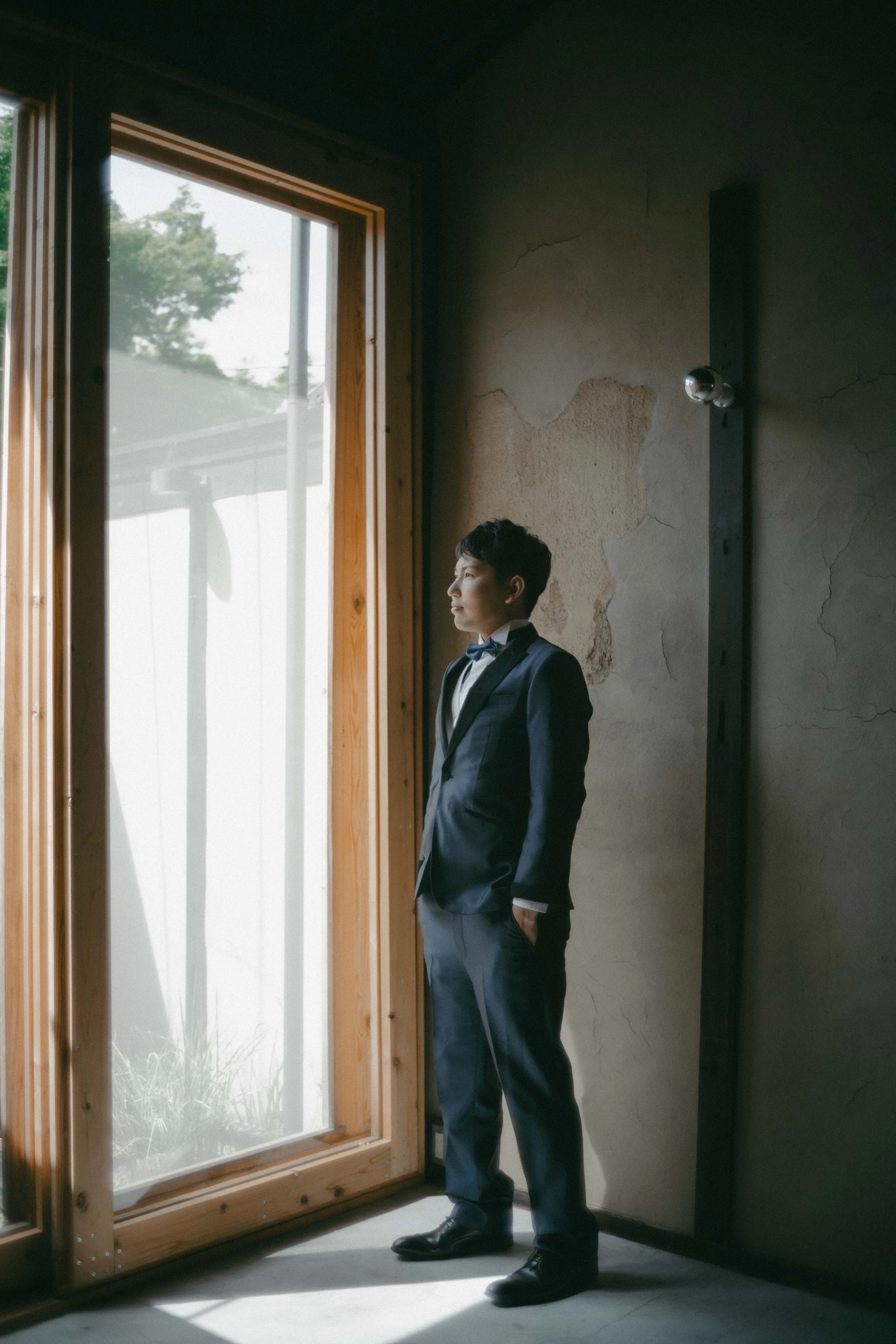A young man in a formal suit and bow tie stands by a large window, looking outside. The room has a rustic, textured wall with exposed patches, and sunlight streams through the window, illuminating his profile.