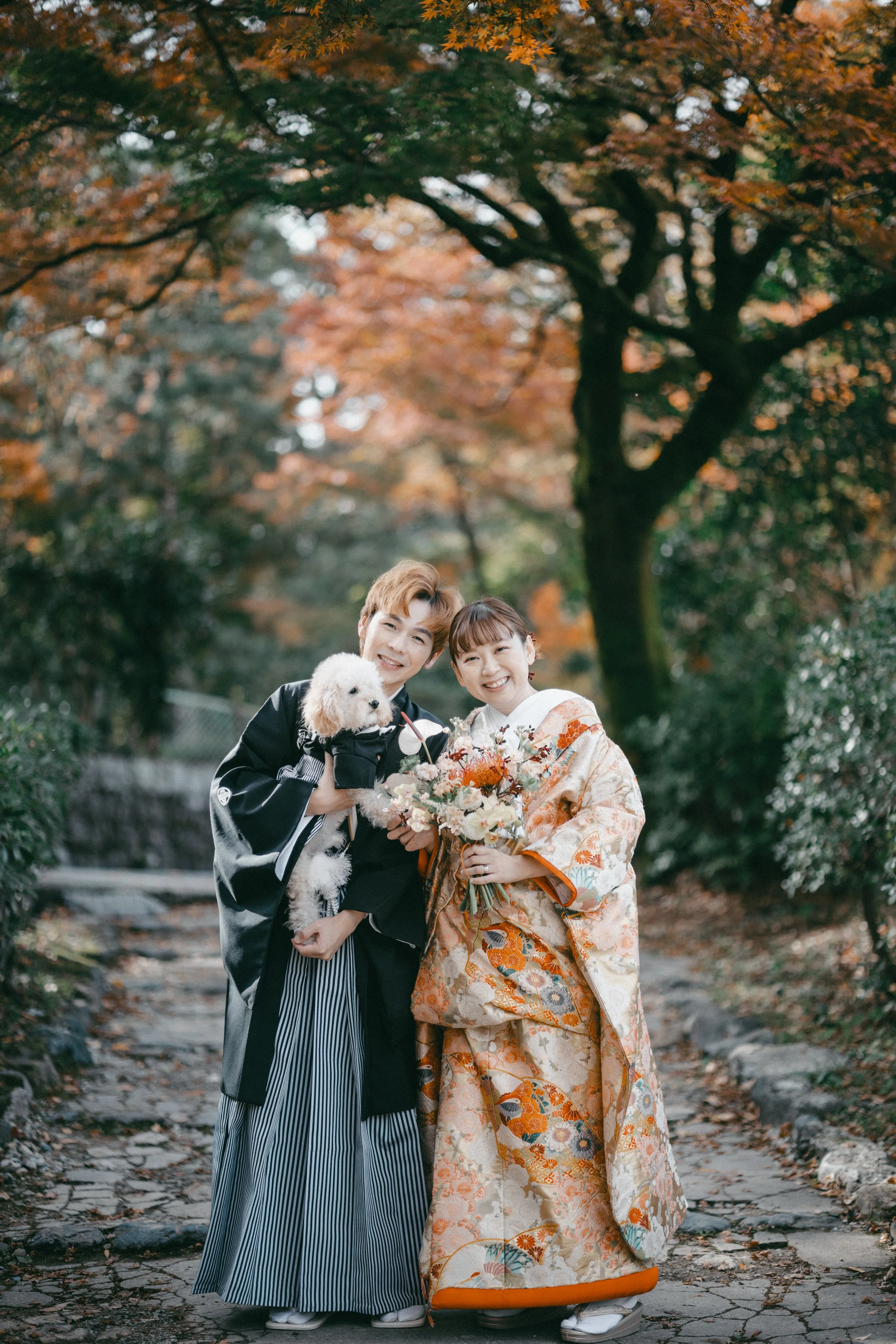 A happy couple in traditional Japanese wedding attire standing outdoors on a stone pathway surrounded by autumn trees, holding a small white dog and a bouquet of flowers.