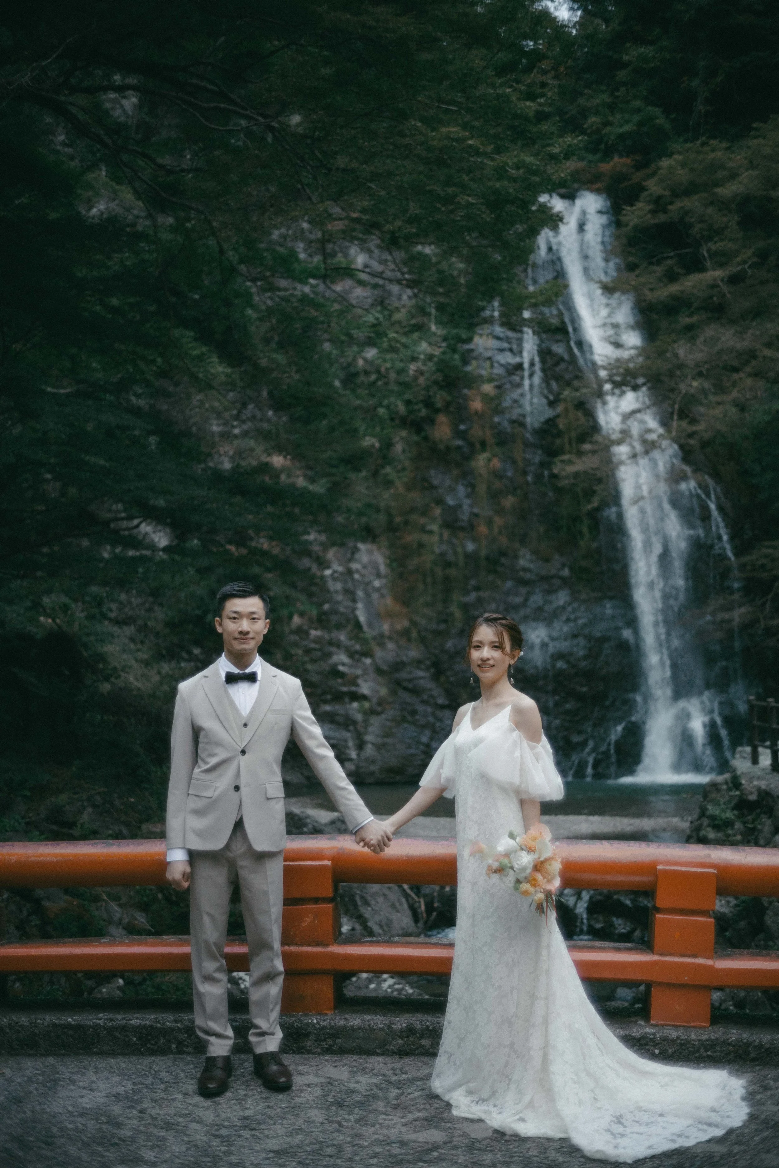 A bride and groom holding hands in front of a waterfall and lush trees.