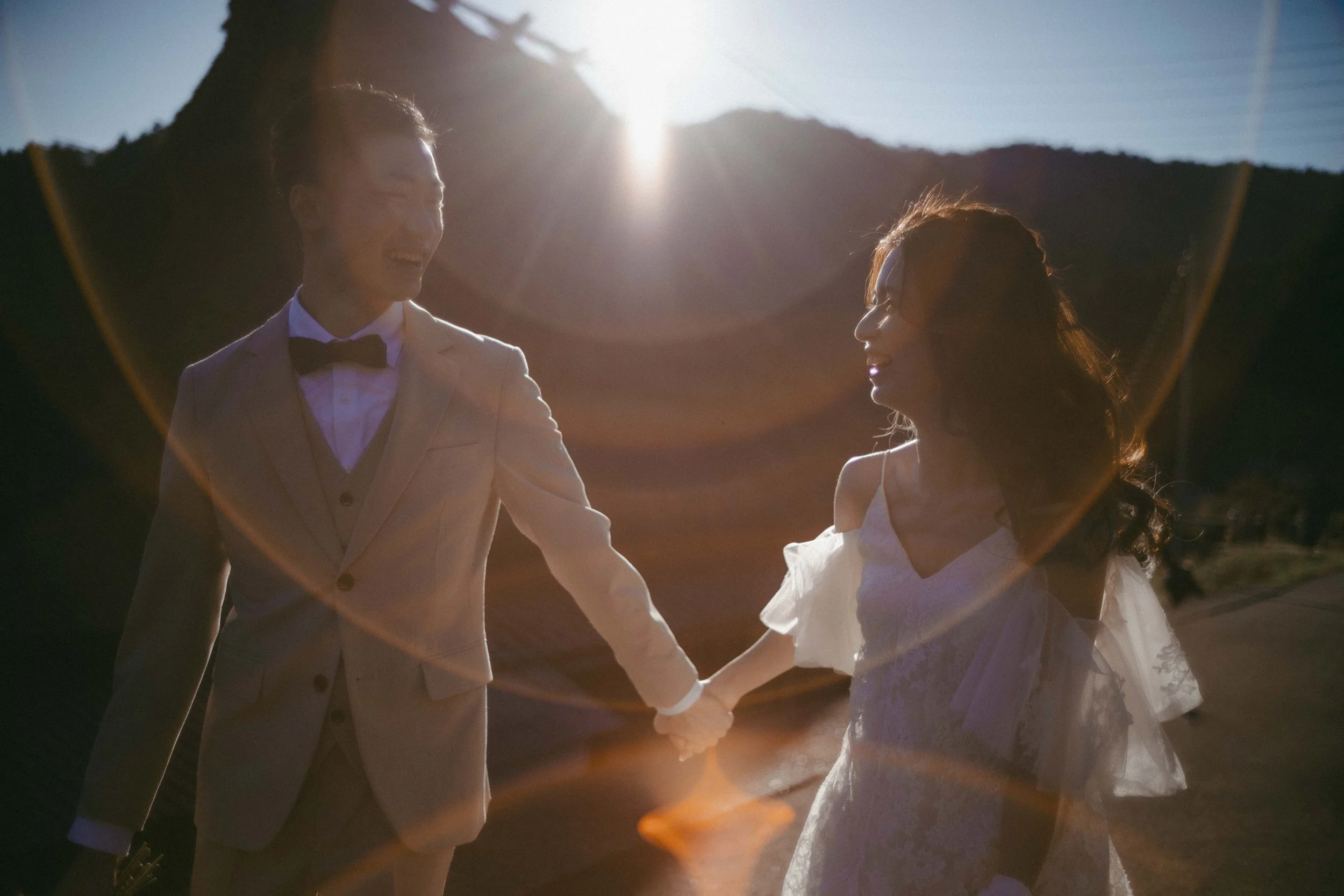 A couple in wedding attire holding hands outdoors at sunset, with sunlight creating a lens flare effect.