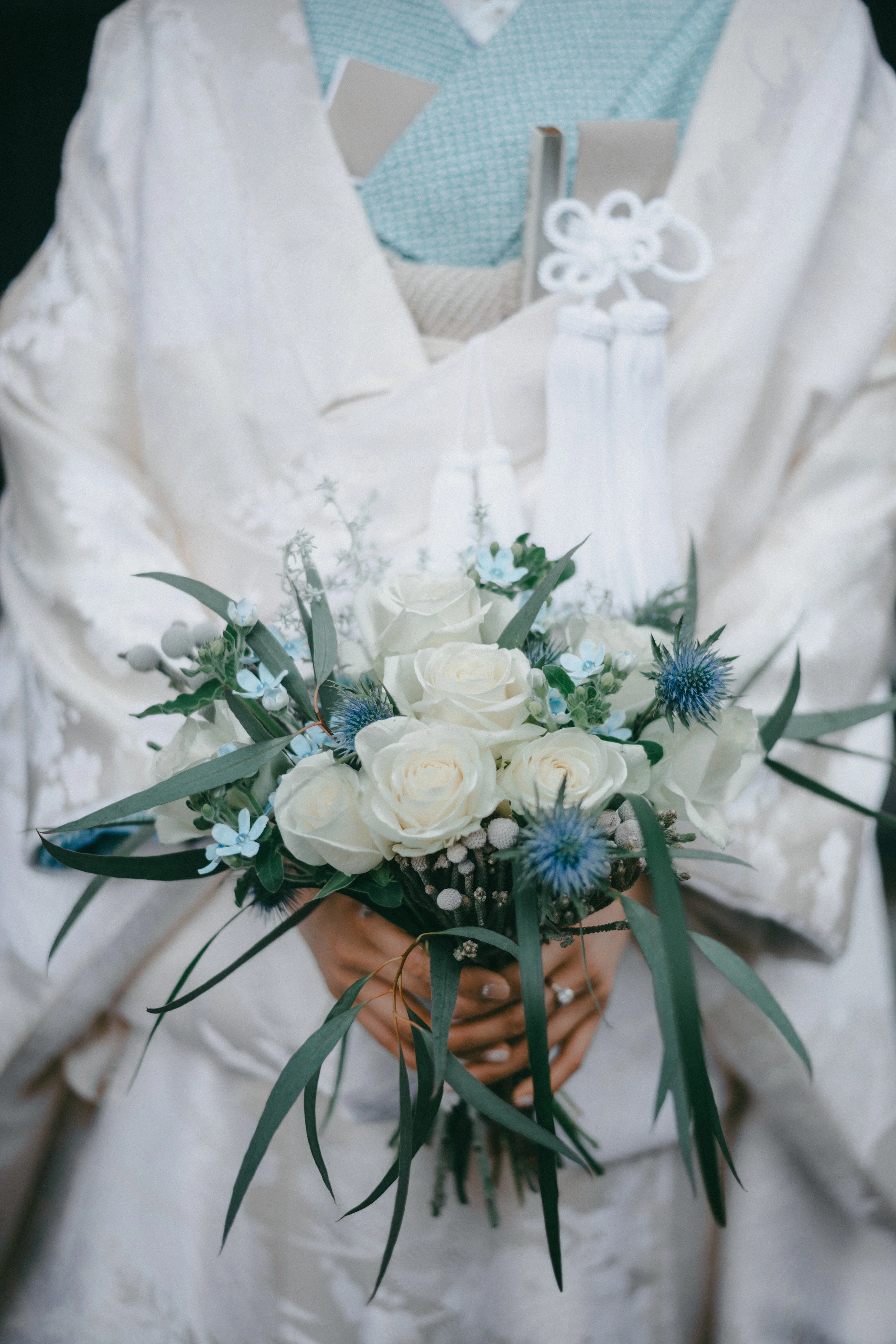 Person holding a bouquet of white roses, blue thistles, and greenery, wearing a white dress with lace details and a white jacket.