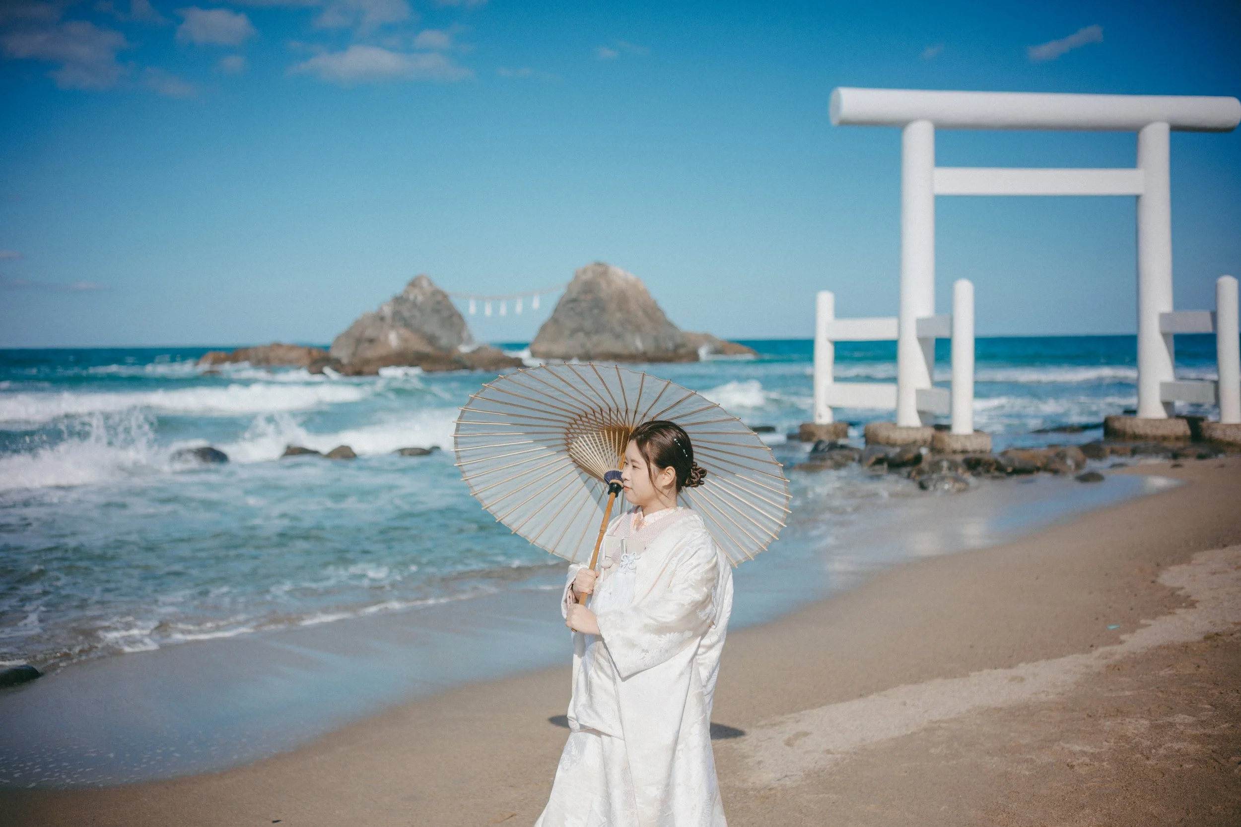 A woman dressed in traditional Japanese attire holding a paper parasol at the beach with a torii gate and rocks in the background.