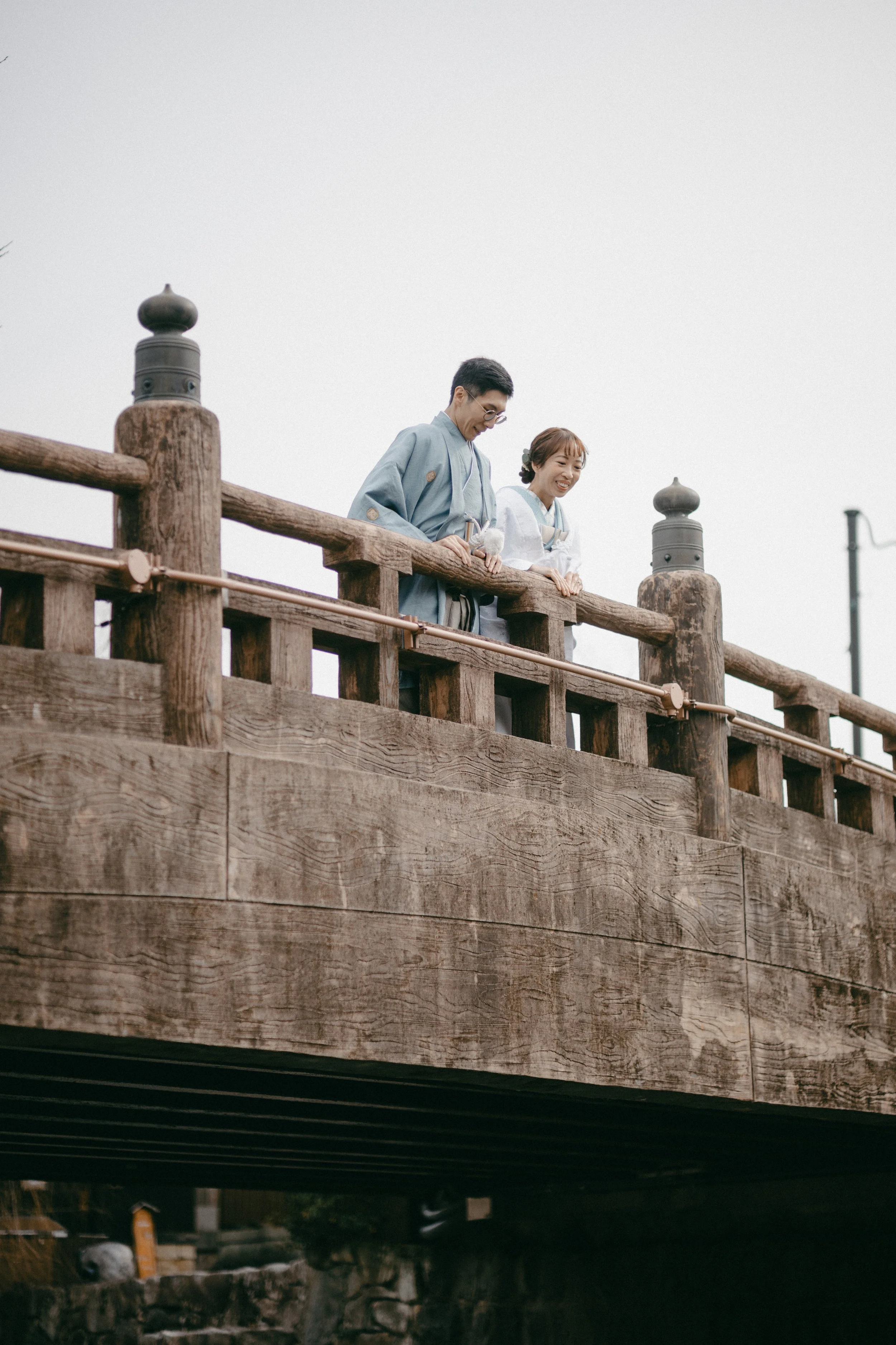 A man and woman in traditional Japanese clothing stand on a wooden bridge, smiling and looking down.