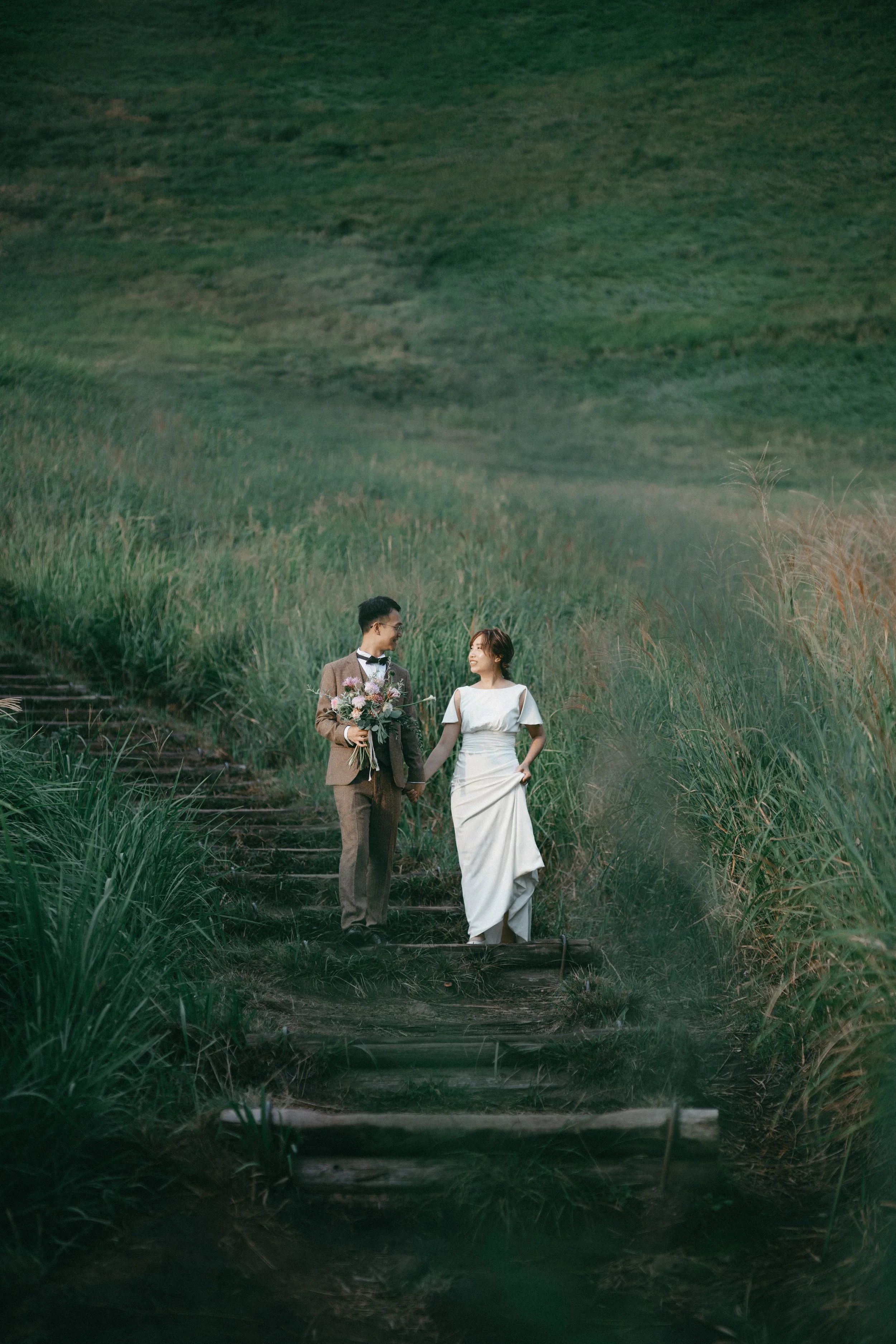 A couple dressed in wedding attire standing on a wooden step path in a green grassy field, holding hands and looking at each other.