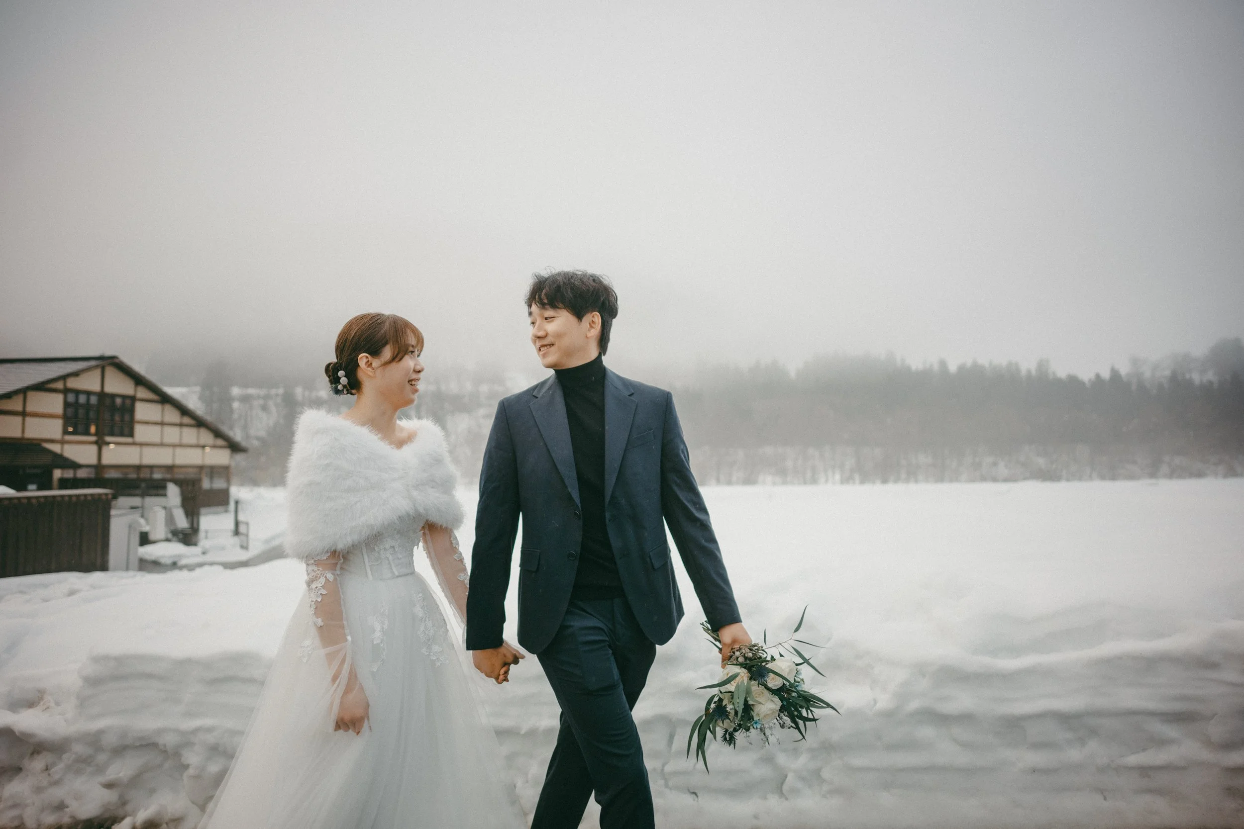 A couple dressed in wedding attire walking hand in hand in a snowy landscape, with a woman in a white wedding dress and a man in a dark suit holding a bouquet. They are smiling at each other against a backdrop of snow-covered ground and cloudy sky.