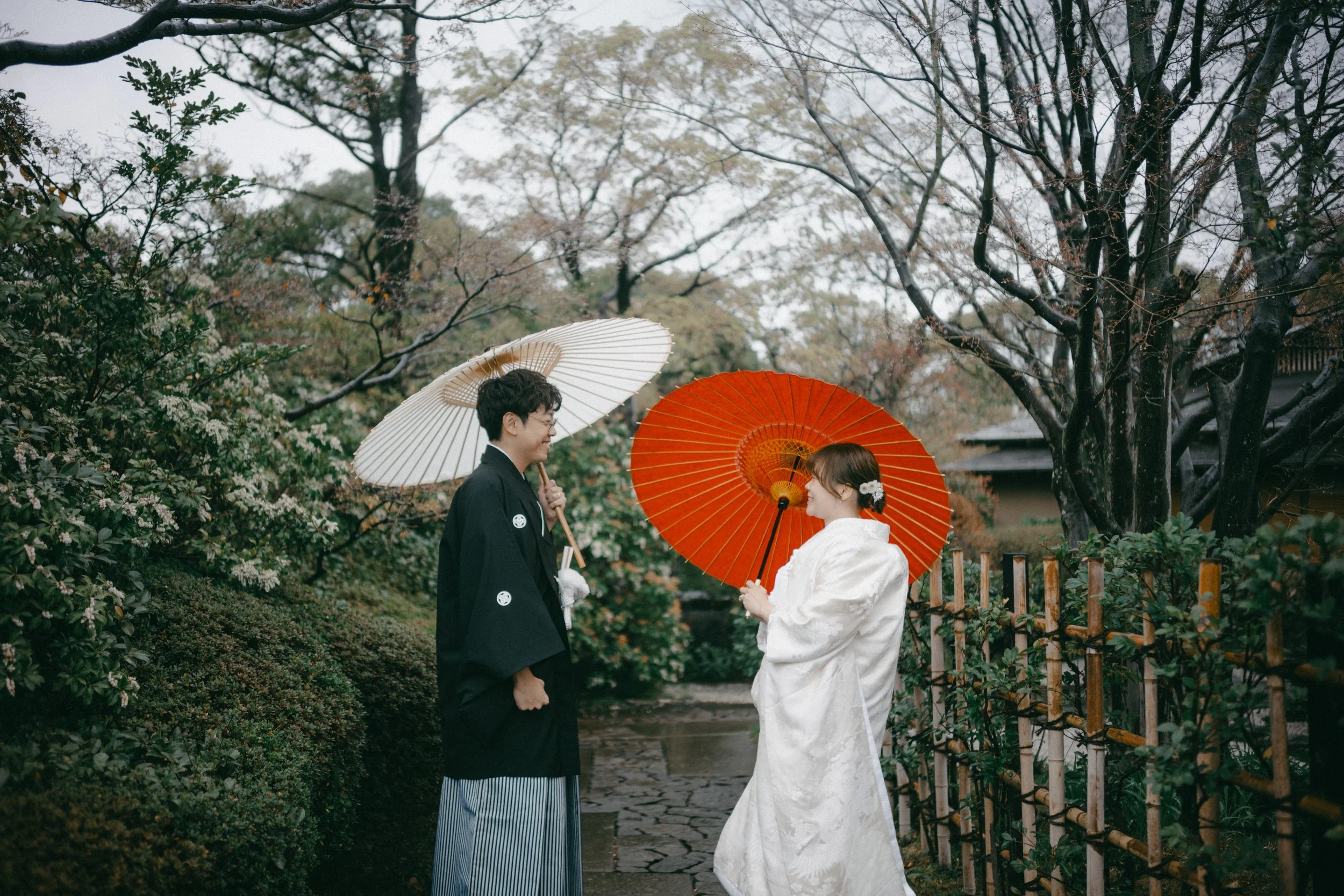 A couple dressed in traditional Japanese wedding attire standing on a stone path outdoors, holding umbrellas, surrounded by trees and lush greenery with overcast sky.