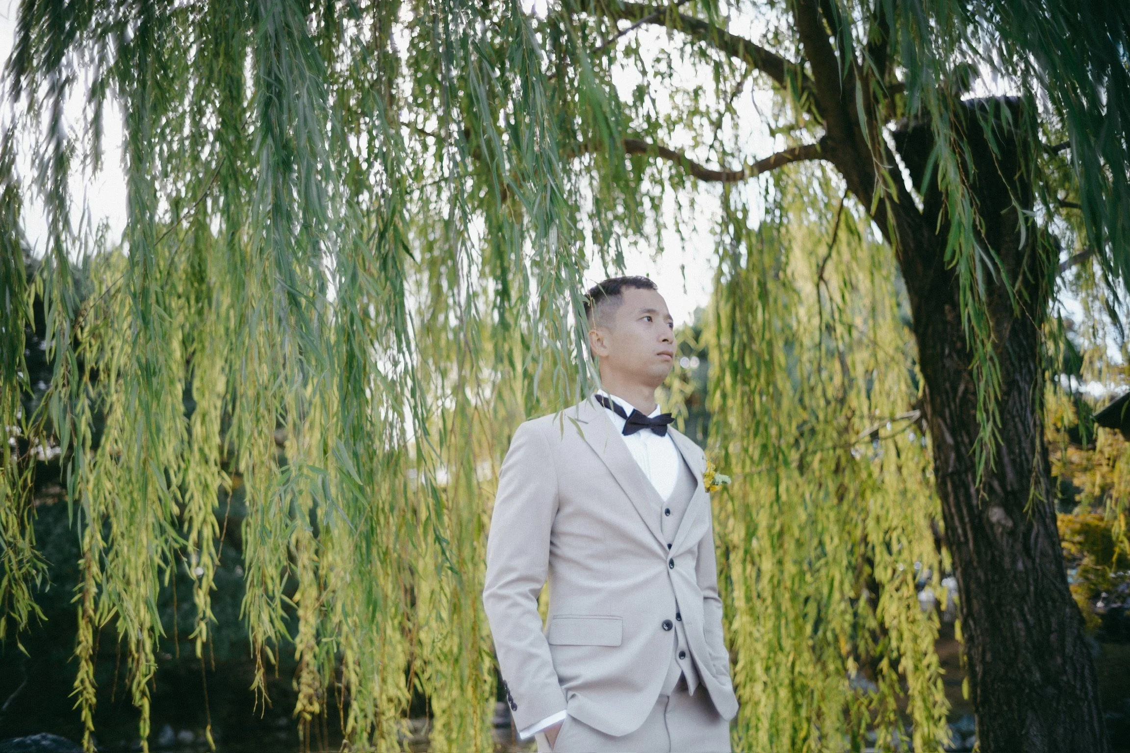Man in a beige suit with a black bowtie standing outdoors under a willow tree.