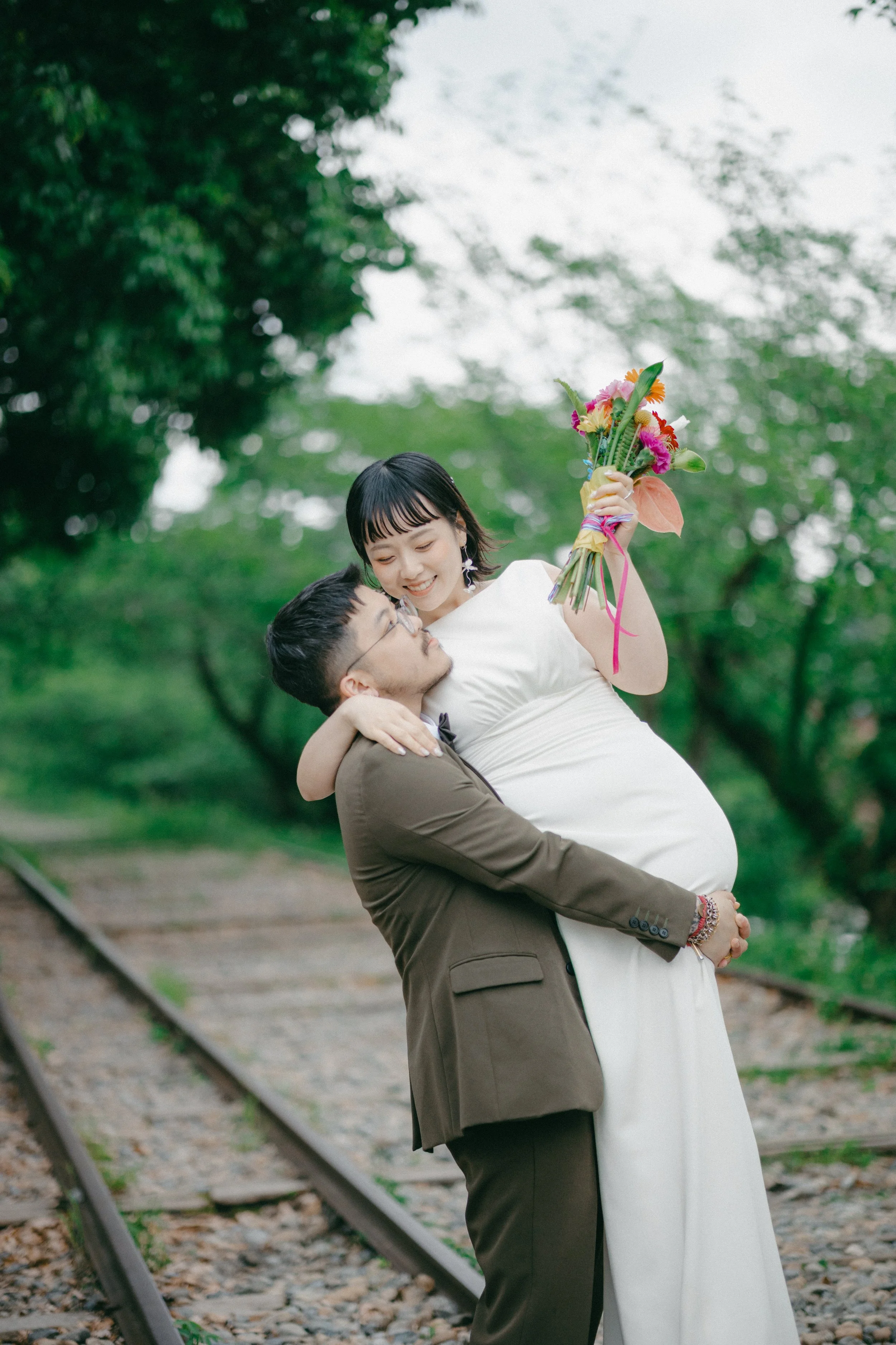 A man in a brown suit lifts a woman in a white dress, holding her close while she holds a colorful bouquet of flowers outdoors on a railroad track surrounded by trees.