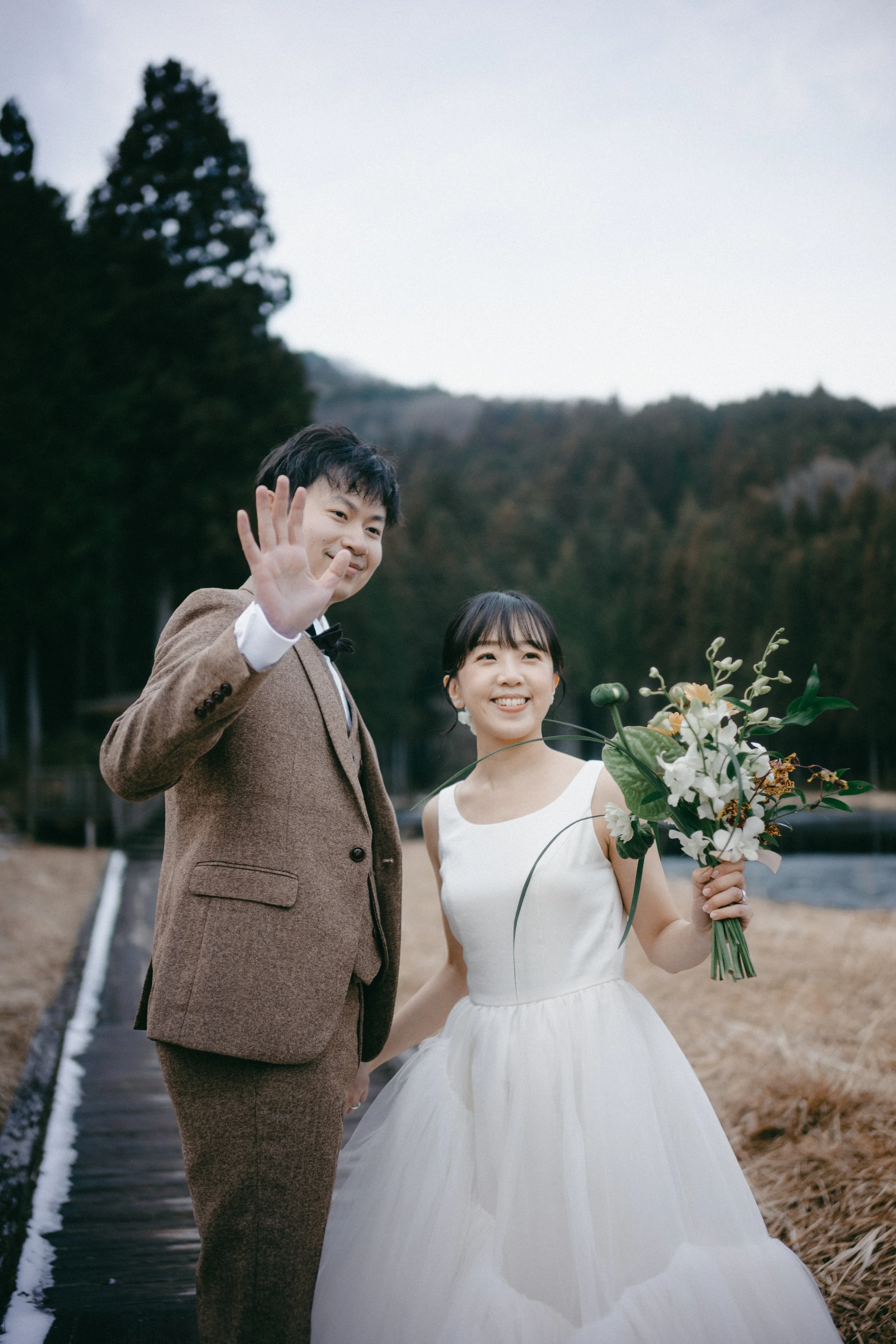A happy couple on their wedding day, holding hands outdoors with a scenic background of trees and mountains, the woman in a white wedding gown holding a bouquet, and the man in a brown suit waving.