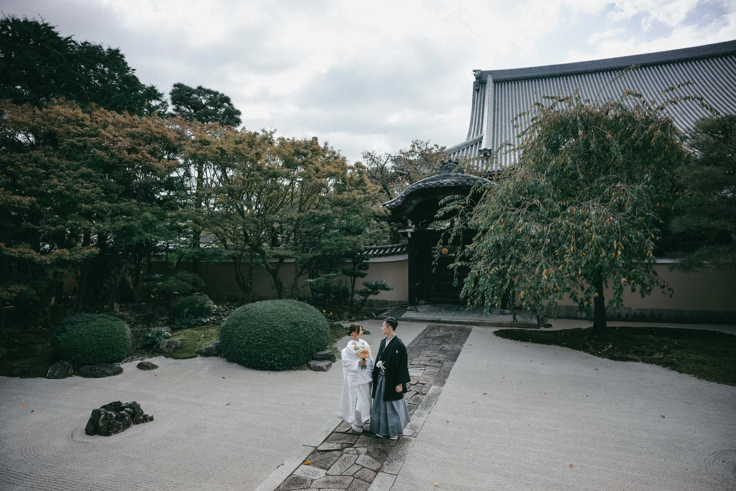 Japanese couple dressed in traditional wedding attire walking in a Japanese garden with trees and a building with a tiled roof in the background.