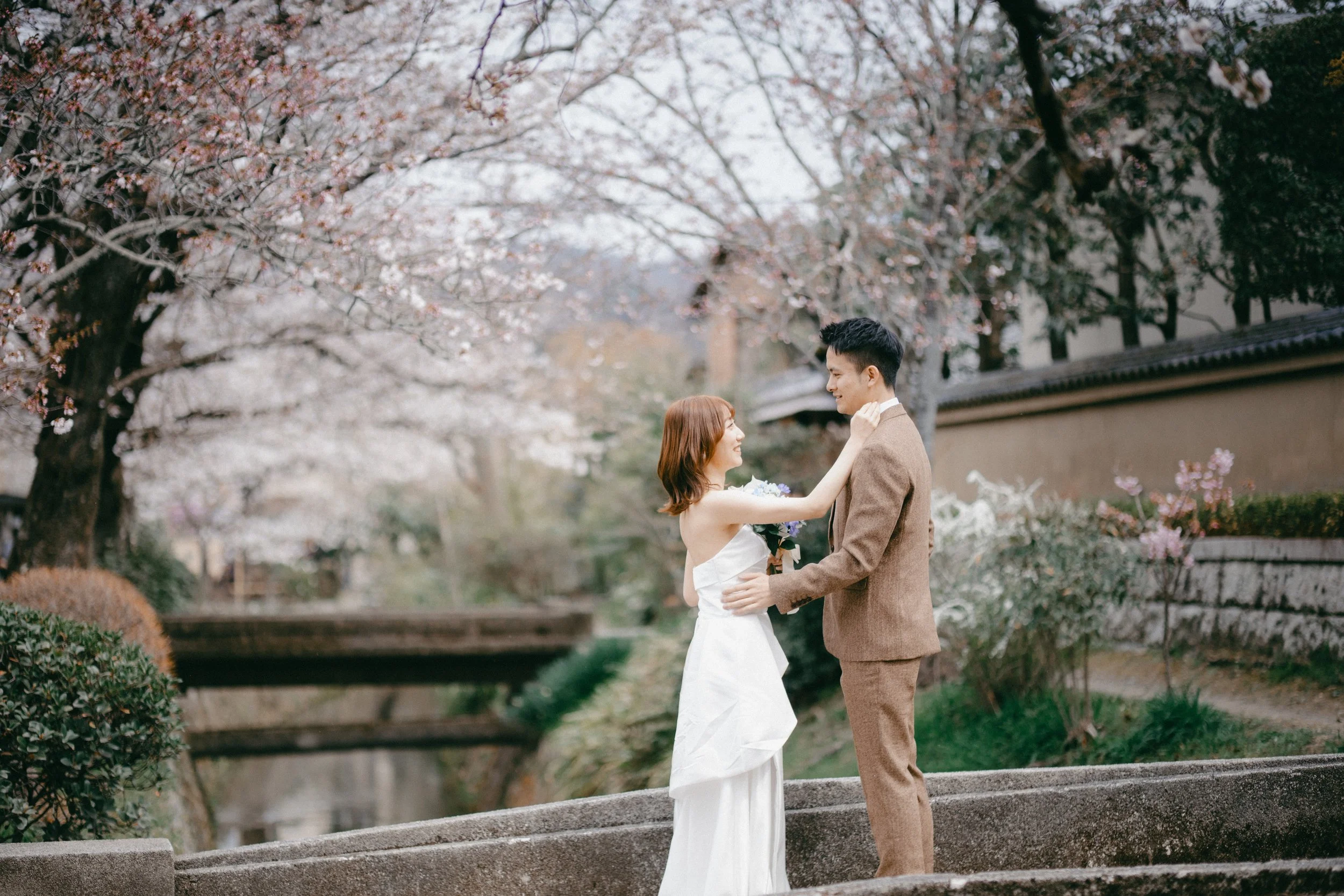 A bride and groom stand facing each other outdoors during spring, with cherry blossom trees in full bloom behind them. The bride is in a white wedding gown holding a bouquet, and the groom is in a brown suit. They are smiling at each other, with the 