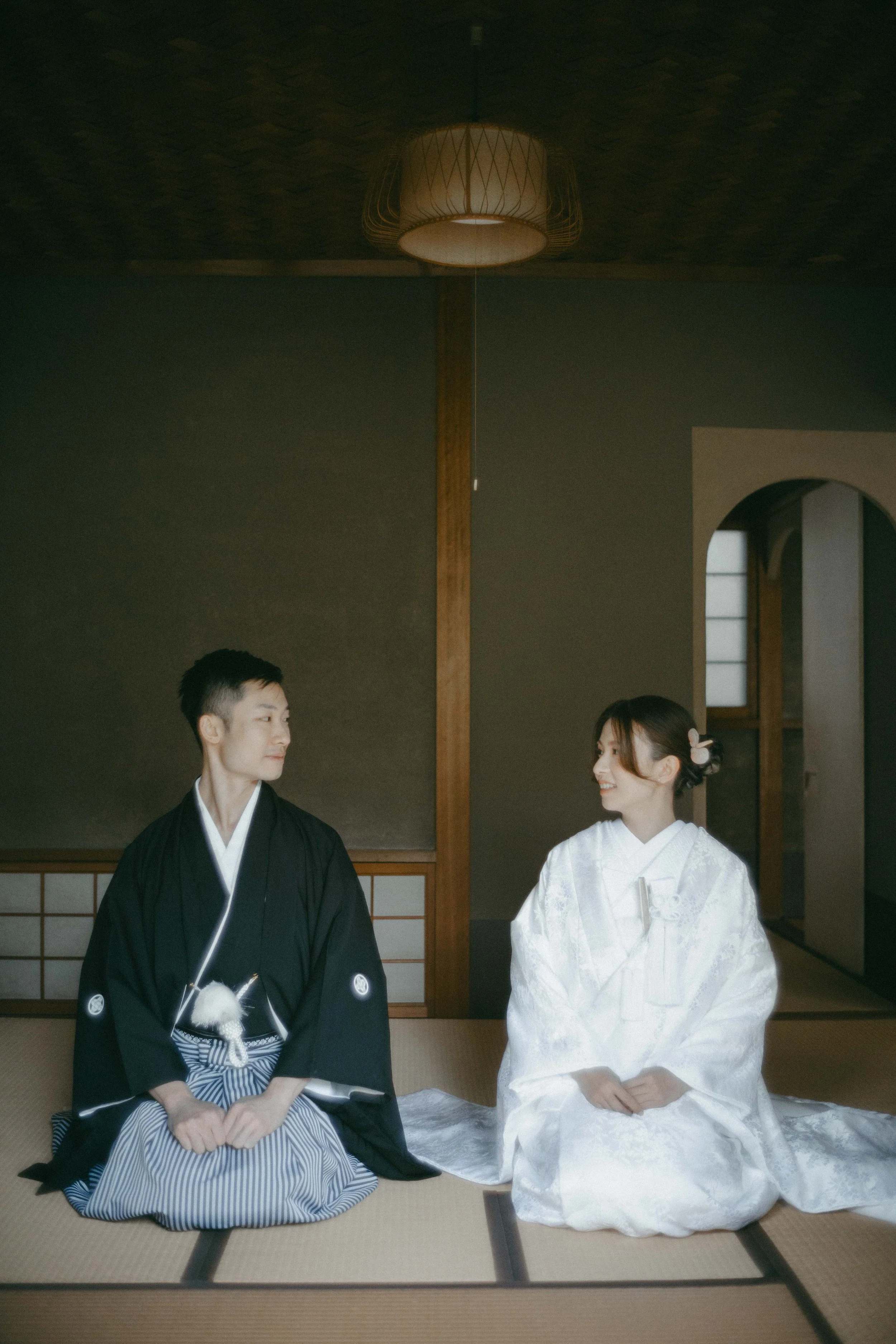 A Japanese man and woman dressed in traditional wedding attire sitting on tatami mats in a room, smiling at each other.