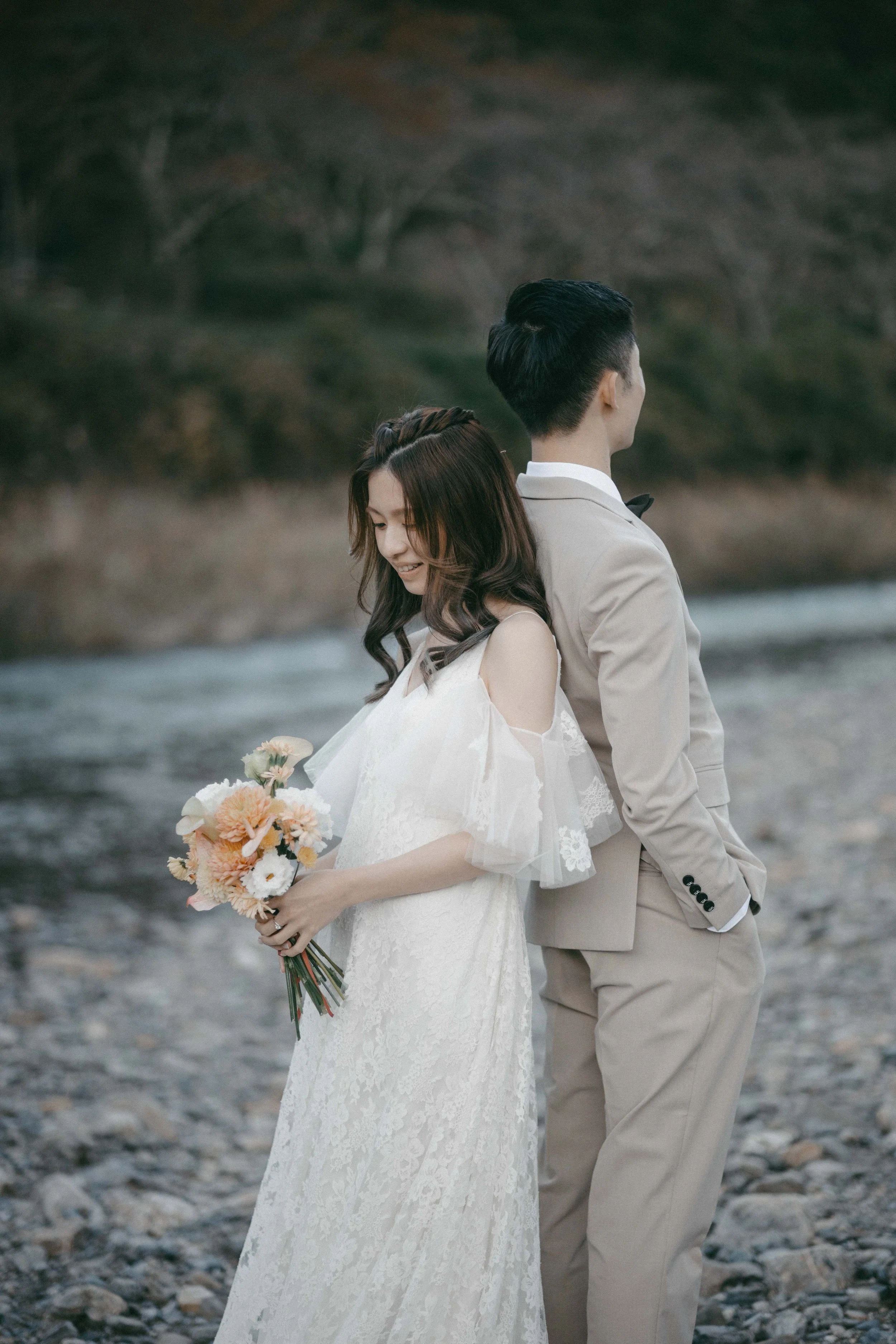 A bride in a white lace wedding dress holding a bouquet of flowers stands with a groom in a beige suit on a rocky riverside; they are back to back with the man looking away and the woman smiling.