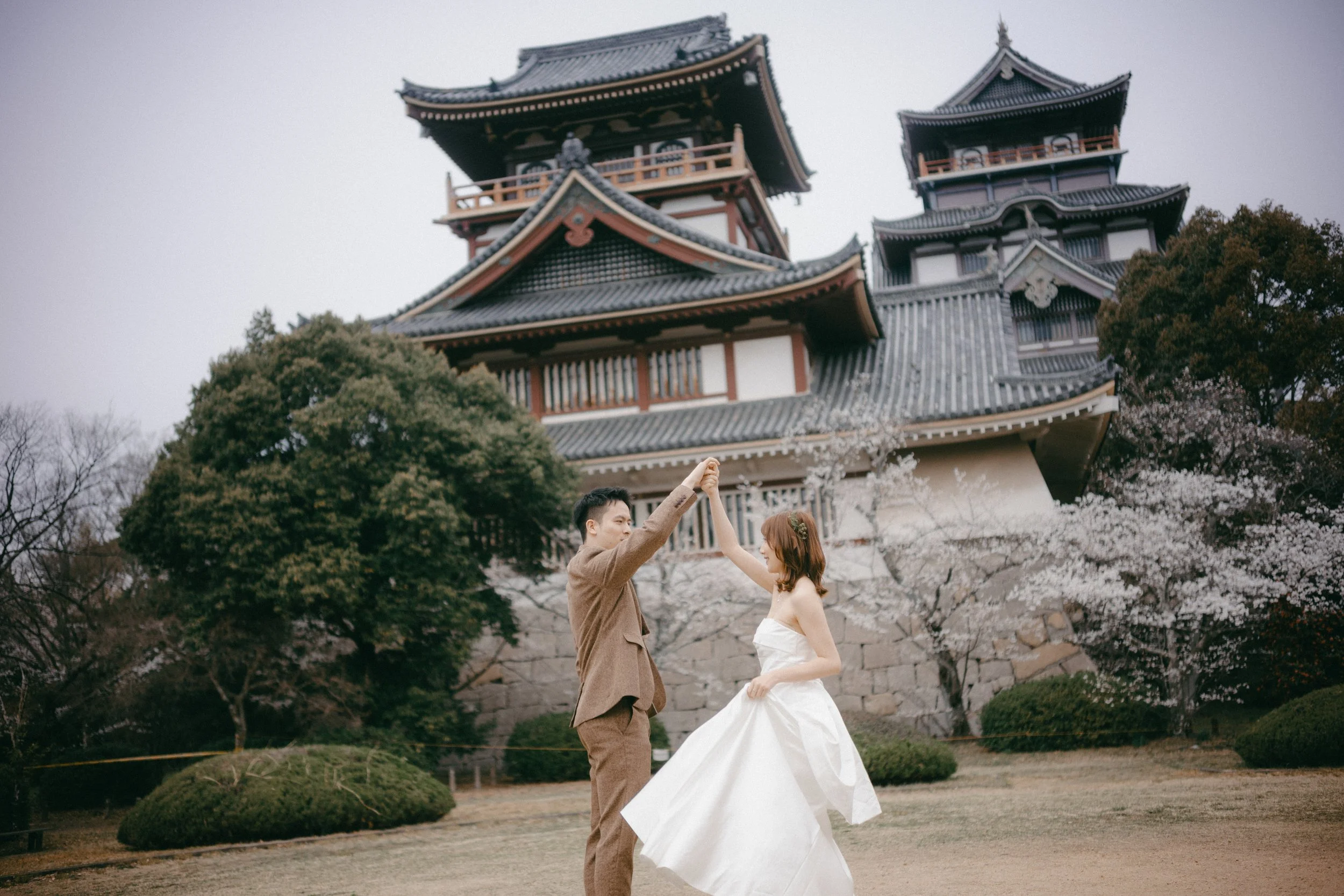 A couple dressed in wedding attire dancing in front of a traditional Japanese castle surrounded by cherry blossom trees.