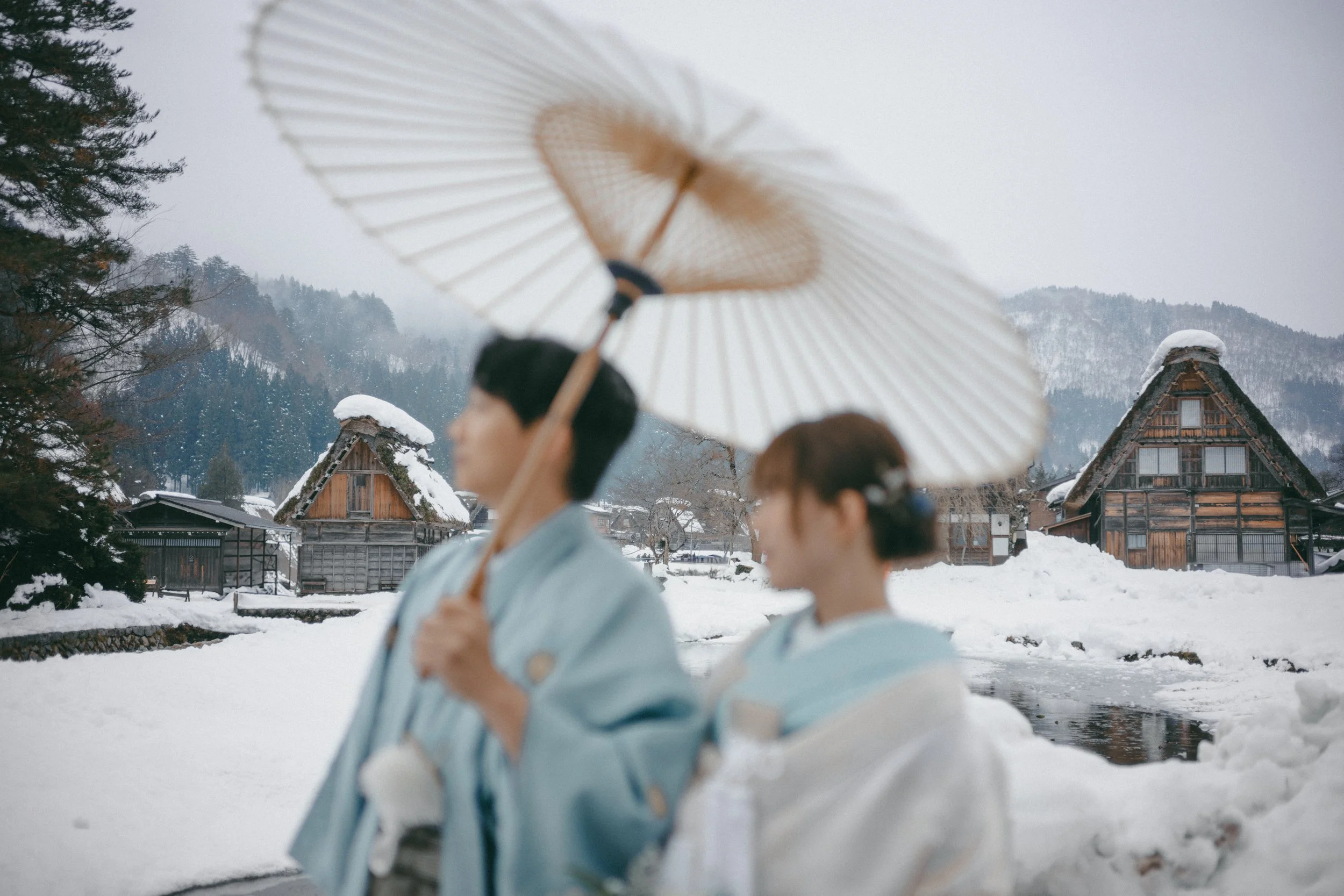 Two women in traditional Japanese kimonos walking through a snowy village with wooden houses, holding umbrellas.