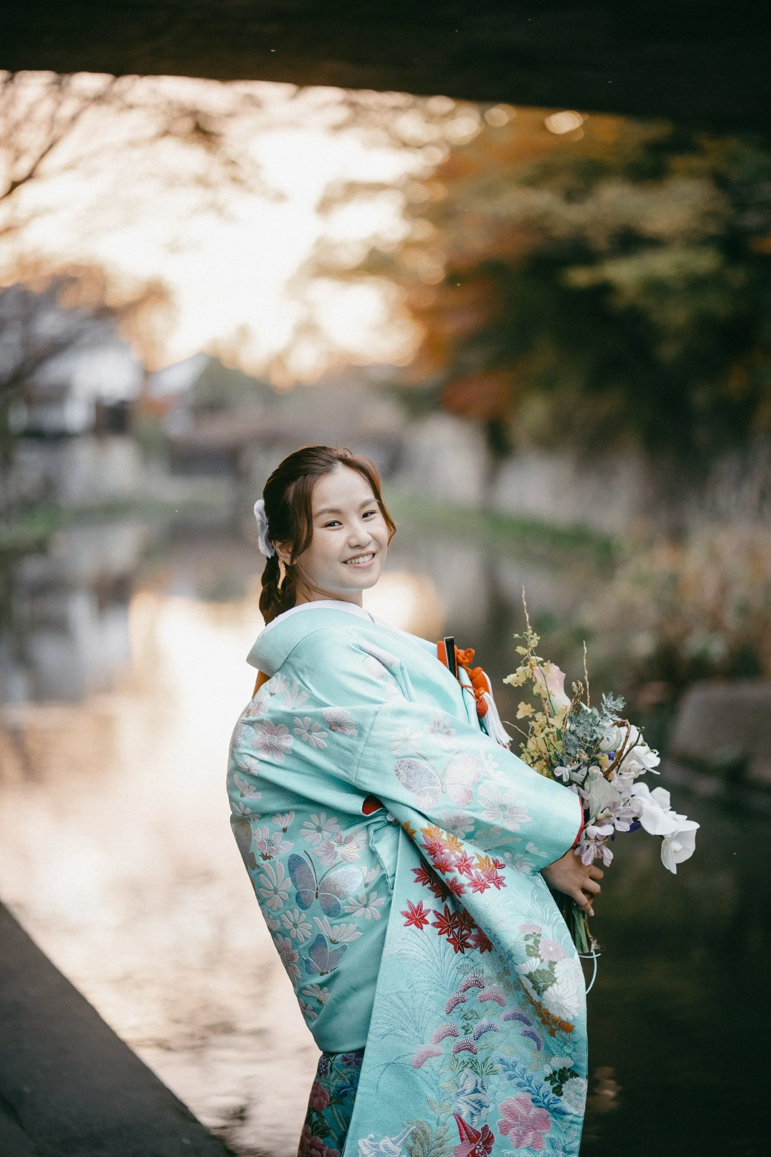 A young woman in a light blue kimono with floral and butterfly patterns, holding a bouquet of flowers, standing by a river at sunset, smiling.