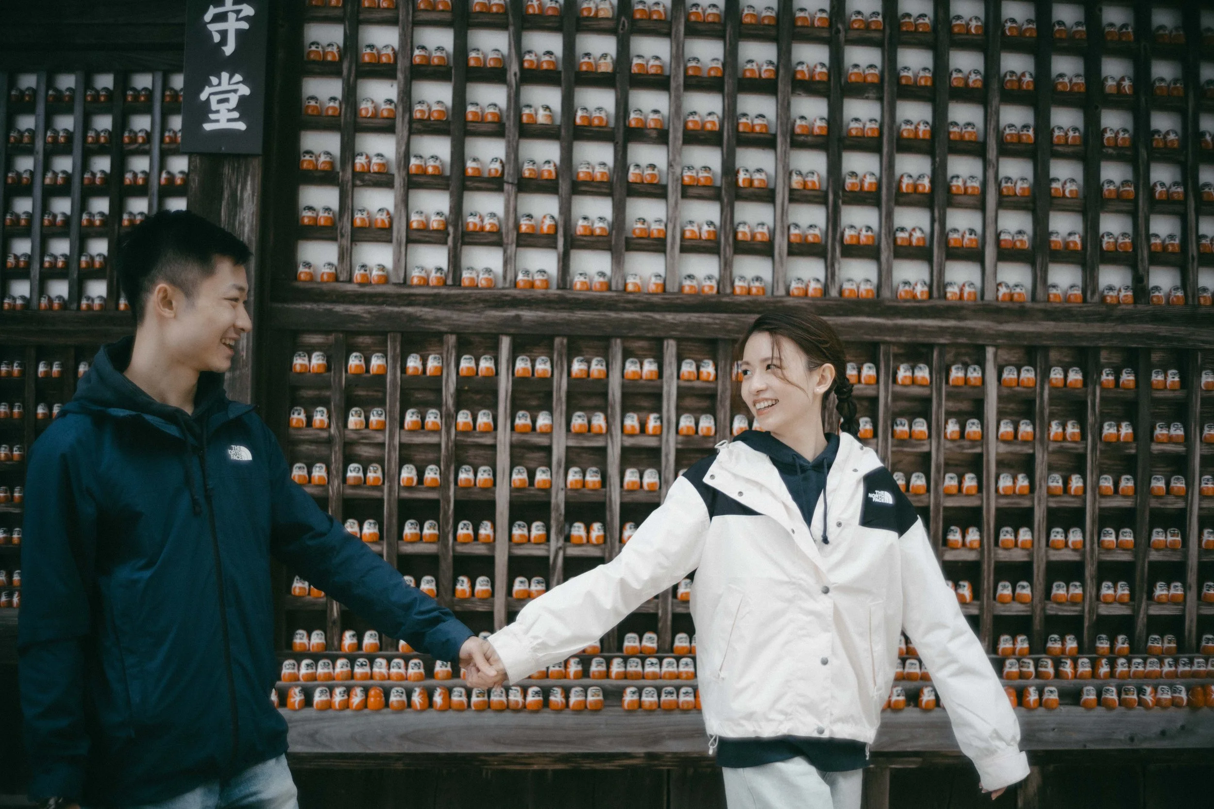 A young man and woman holding hands and smiling at each other in front of a large wooden wall filled with small jars, possibly in a temple or traditional setting.