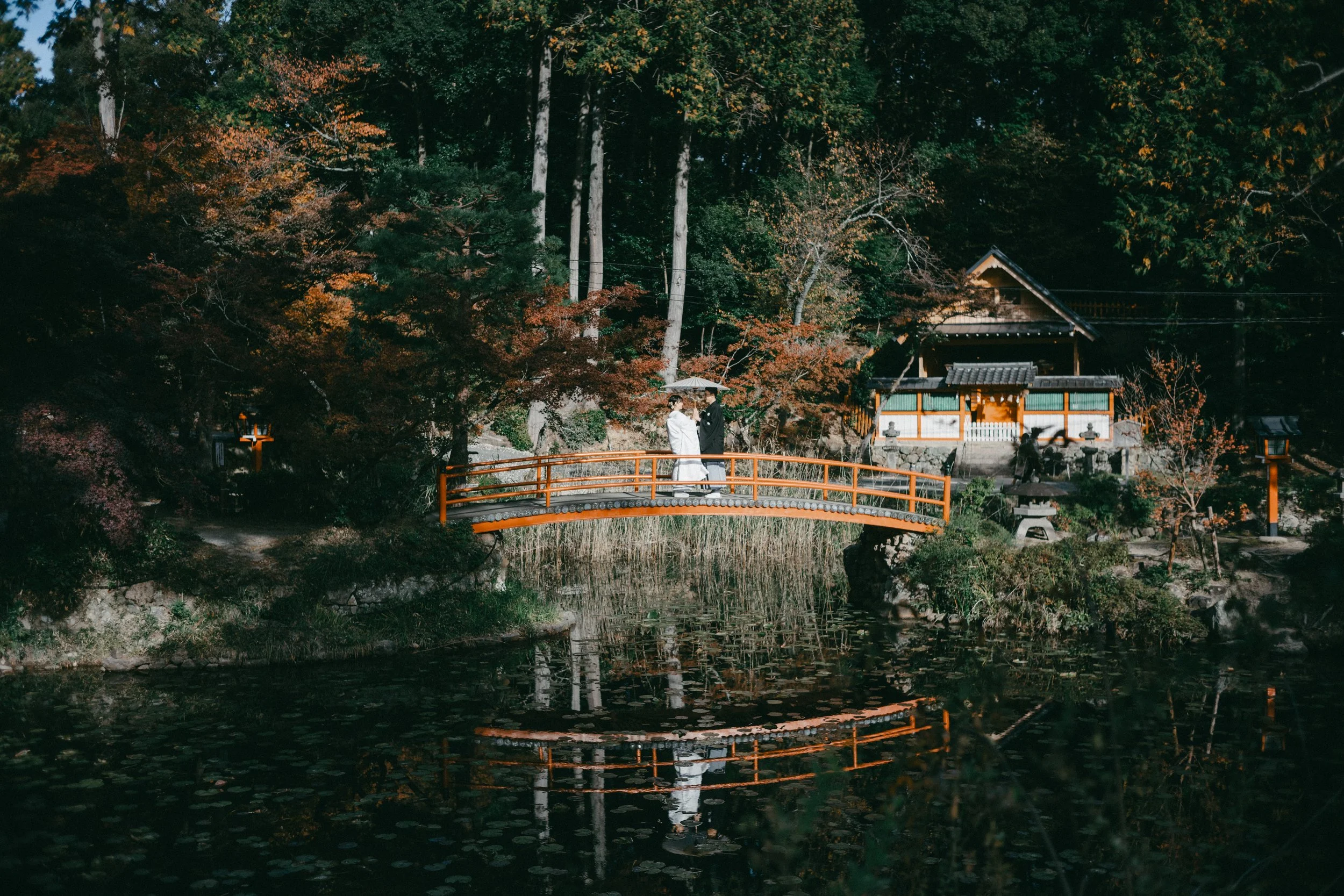 A couple dressed in traditional wedding attire standing on a small orange bridge over a pond, with trees and a traditional Japanese building in the background.