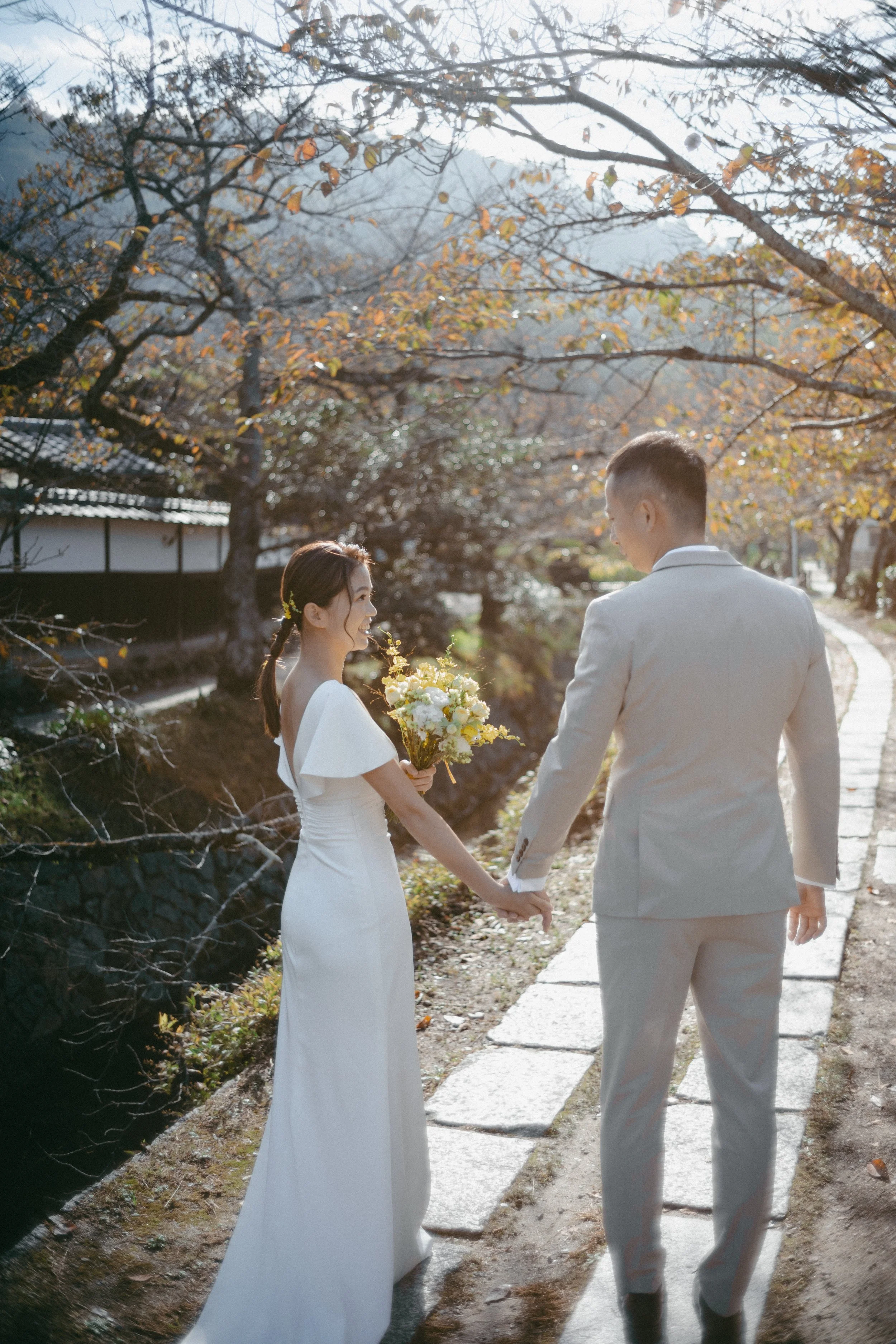 A bride and groom walking hand in hand on a stone path, surrounded by trees with autumn leaves, during late afternoon or early evening.