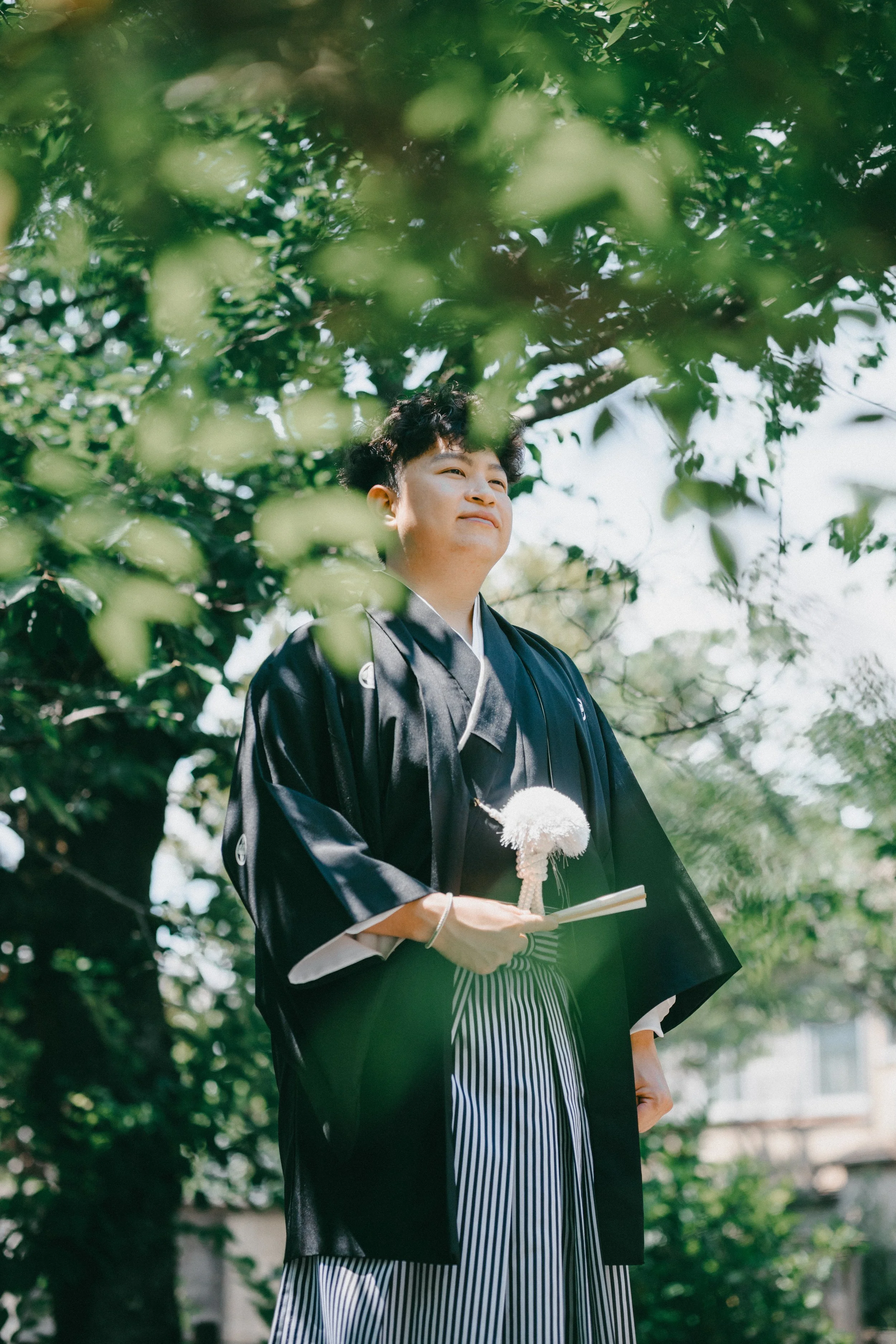 A young man dressed in traditional Japanese attire standing outdoors amidst green foliage, holding a decorative ceremonial object and a paper fan, during a possibly cultural or ceremonial event.