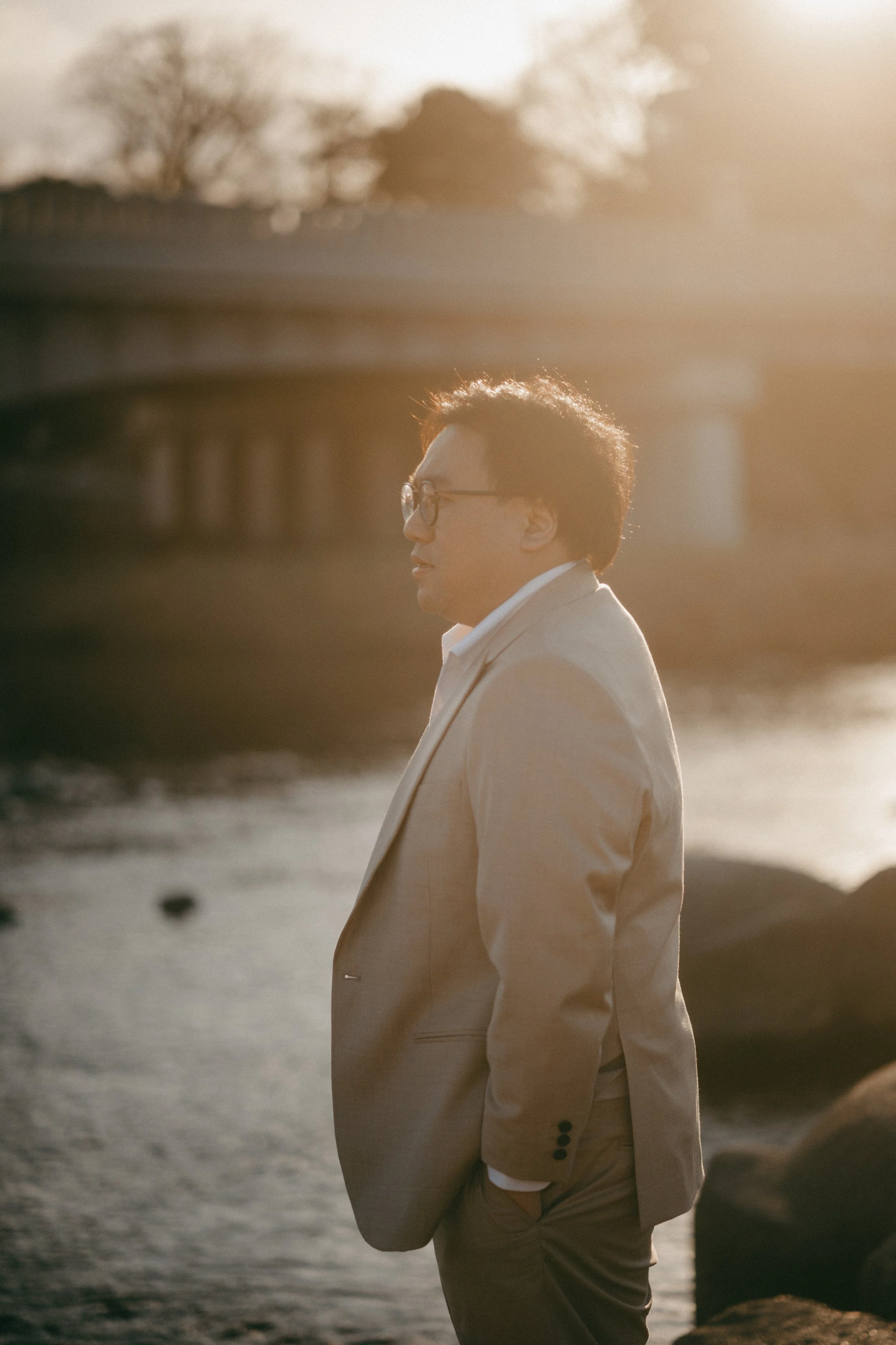 A man wearing glasses in a light-colored suit stands by a river with a bridge in the background during sunset.