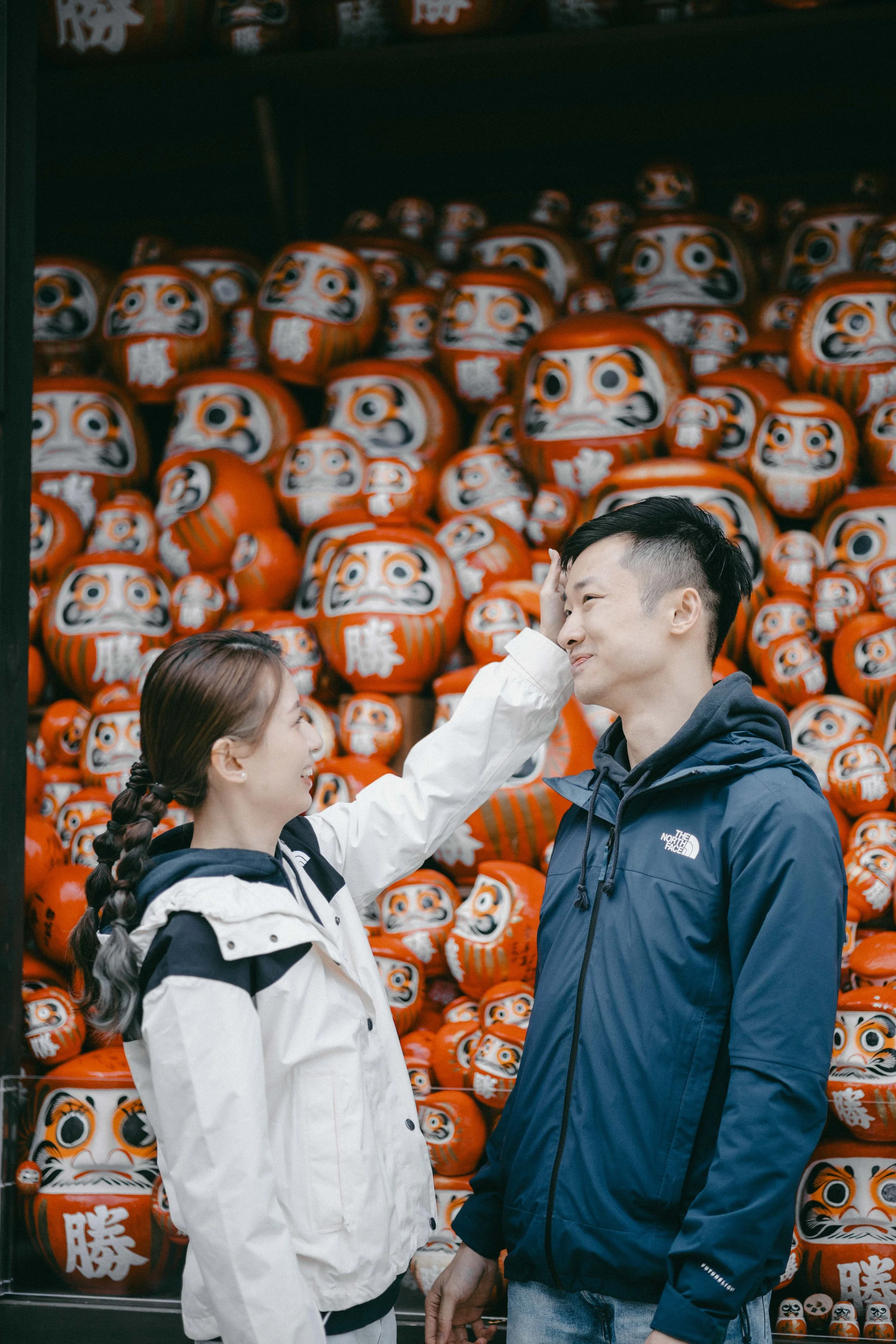 A woman is touching a man's forehead in a playful manner in front of a display of orange Daruma dolls.