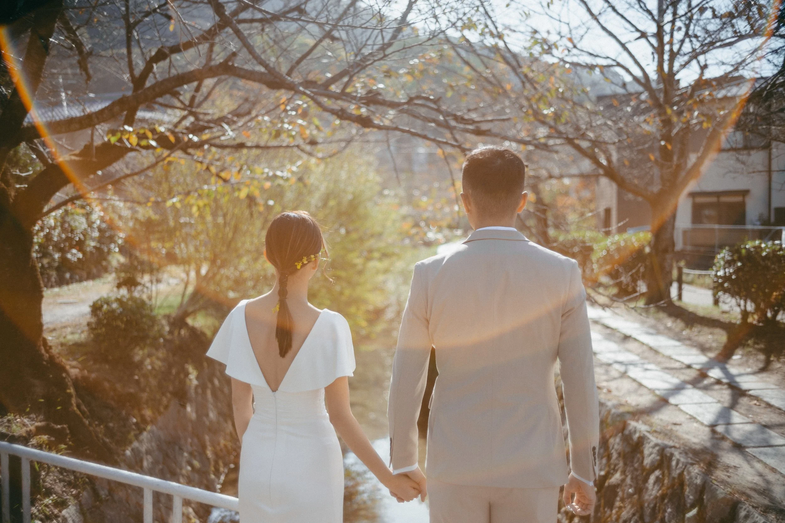 A couple dressed in white holding hands while walking along a small river or canal on a sunny day with trees and buildings in the background.