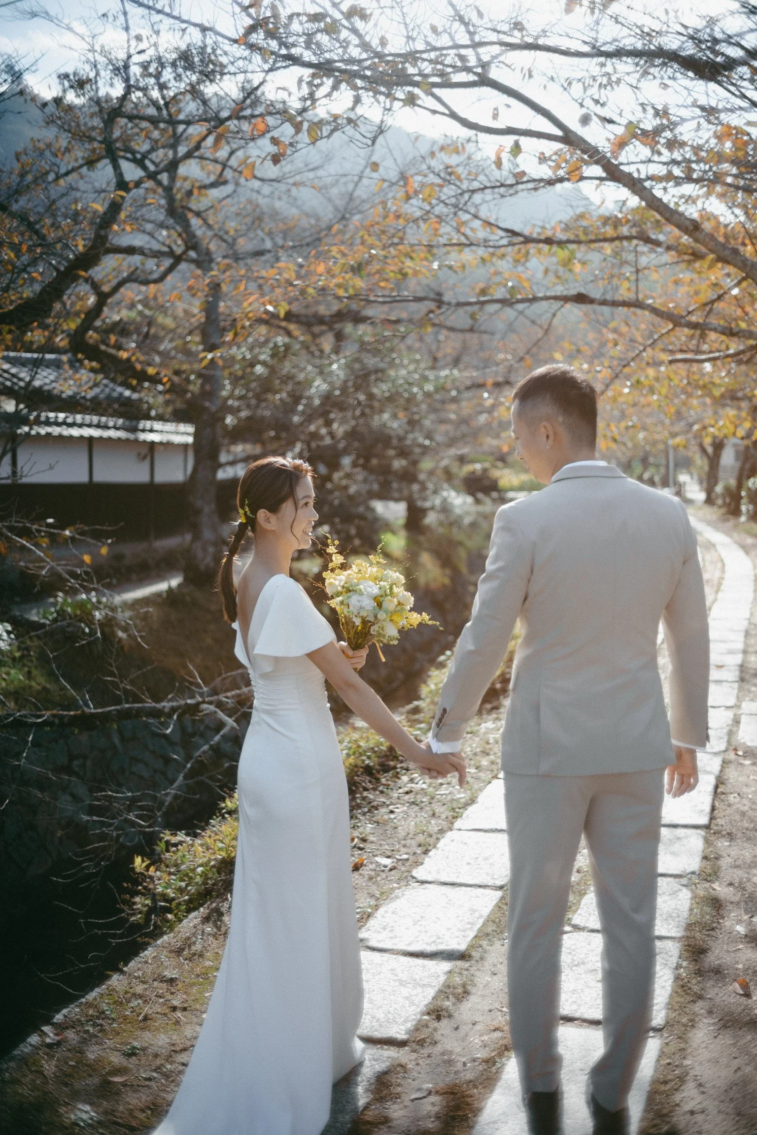 A bride and groom holding hands and walking along a stone path in a park during autumn, with trees with orange and yellow leaves