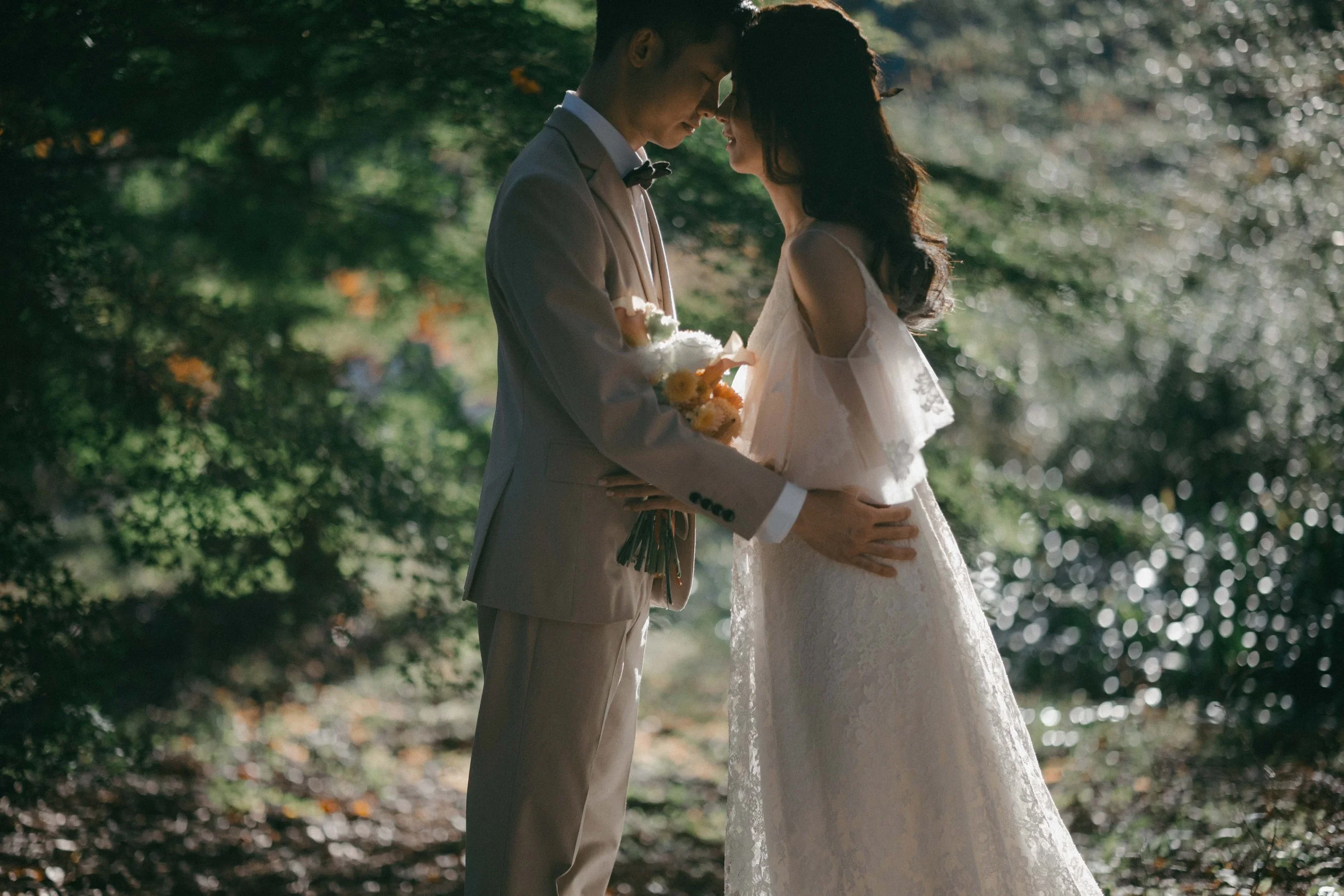 A bride and groom standing close together in a forest, touching foreheads, with the groom holding a bouquet of flowers.