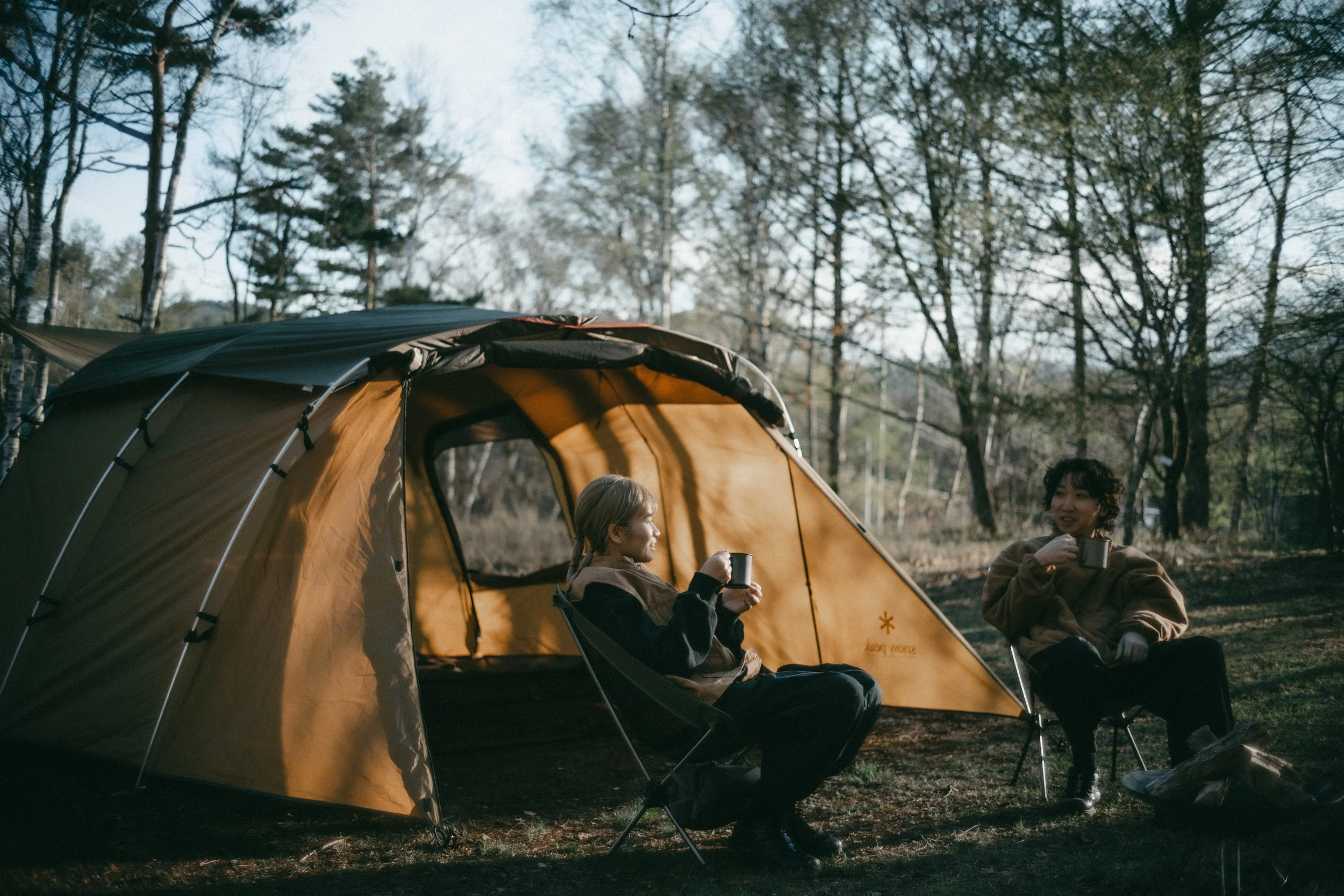 Two women sitting and relaxing outside a yellow camping tent in a wooded area, enjoying warm drinks.