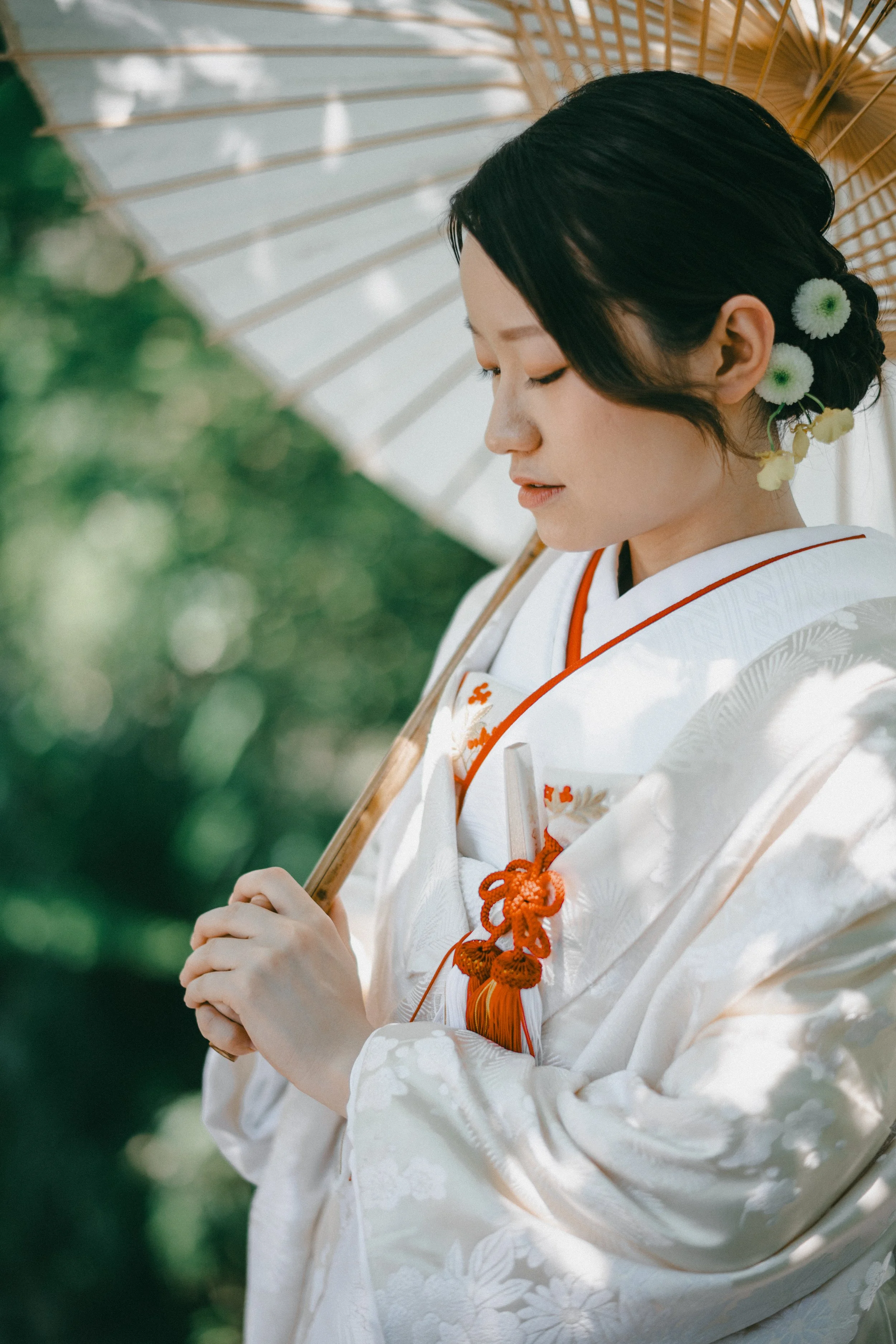 A woman dressed in traditional Japanese wedding attire, holding a parasol. She has flowers in her hair and is standing outdoors with a blurred green background.