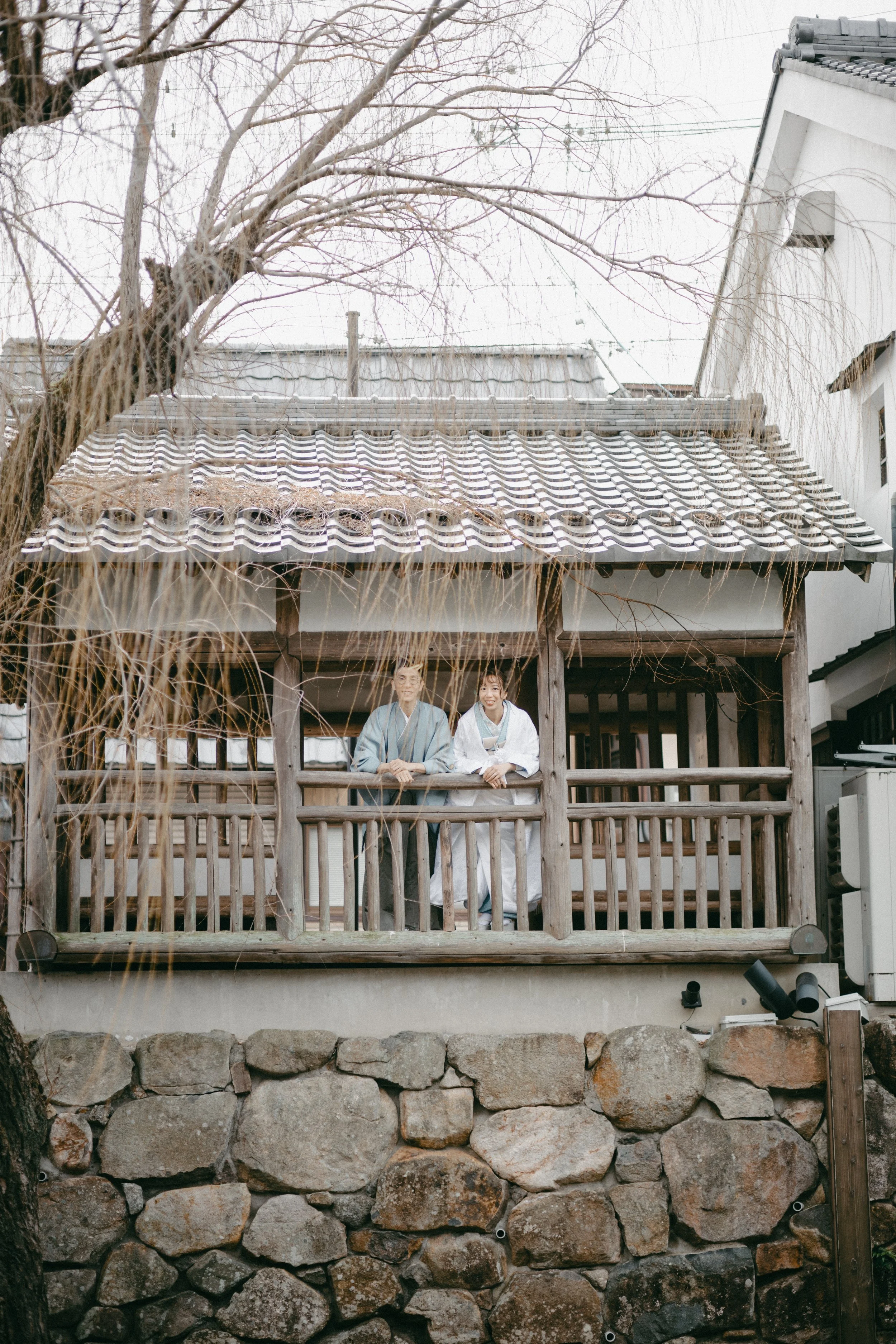Two people dressed in traditional Japanese kimonos standing on a wooden balcony of a small house, with a stone foundation and a tiled roof, in a winter setting with leafless trees.
