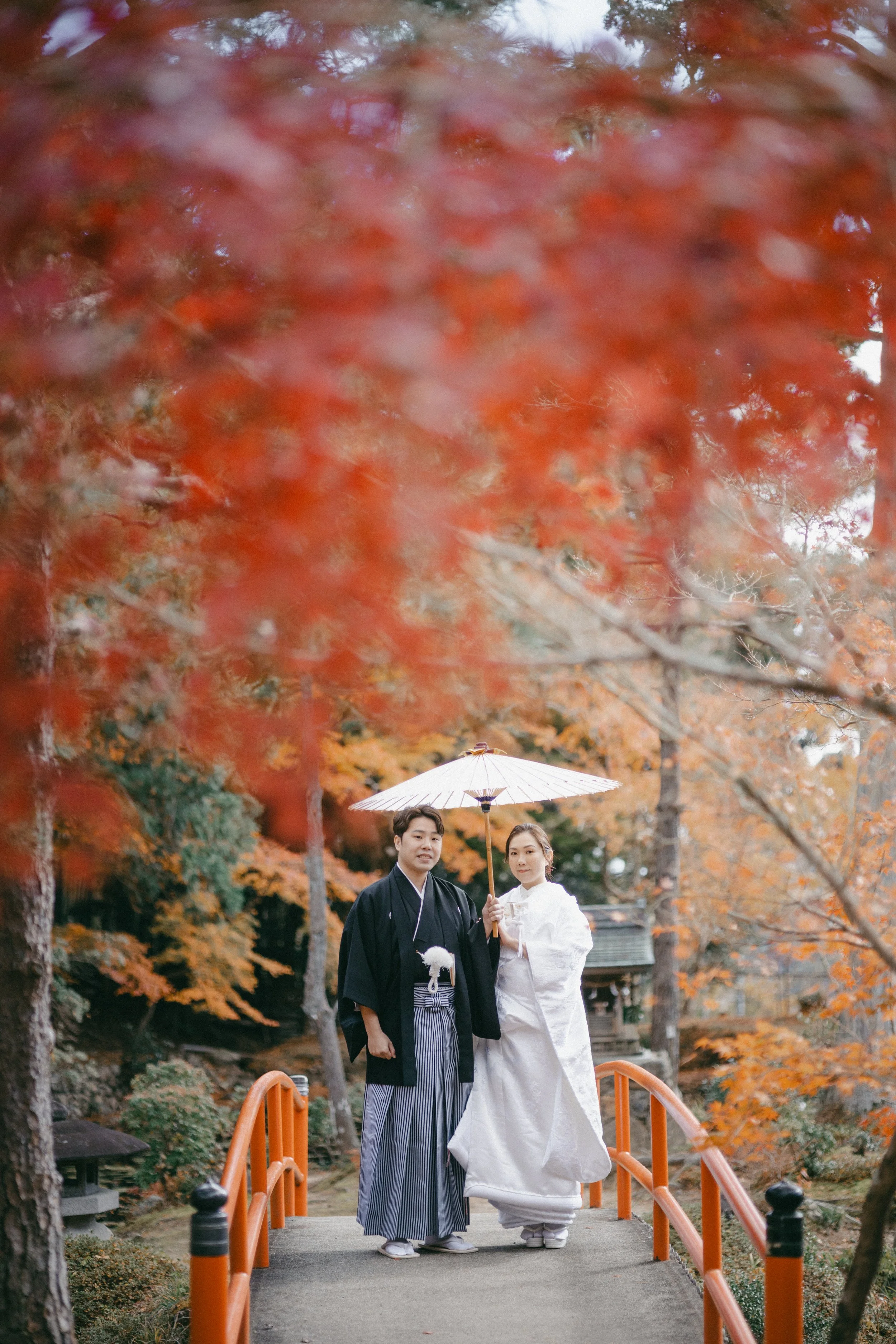 A couple dressed in traditional Japanese wedding attire standing on a small bridge during autumn. The woman is in a white kimono holding a white paper umbrella, and the man is in a black kimono with a striped hakama. They are surrounded by colorful f