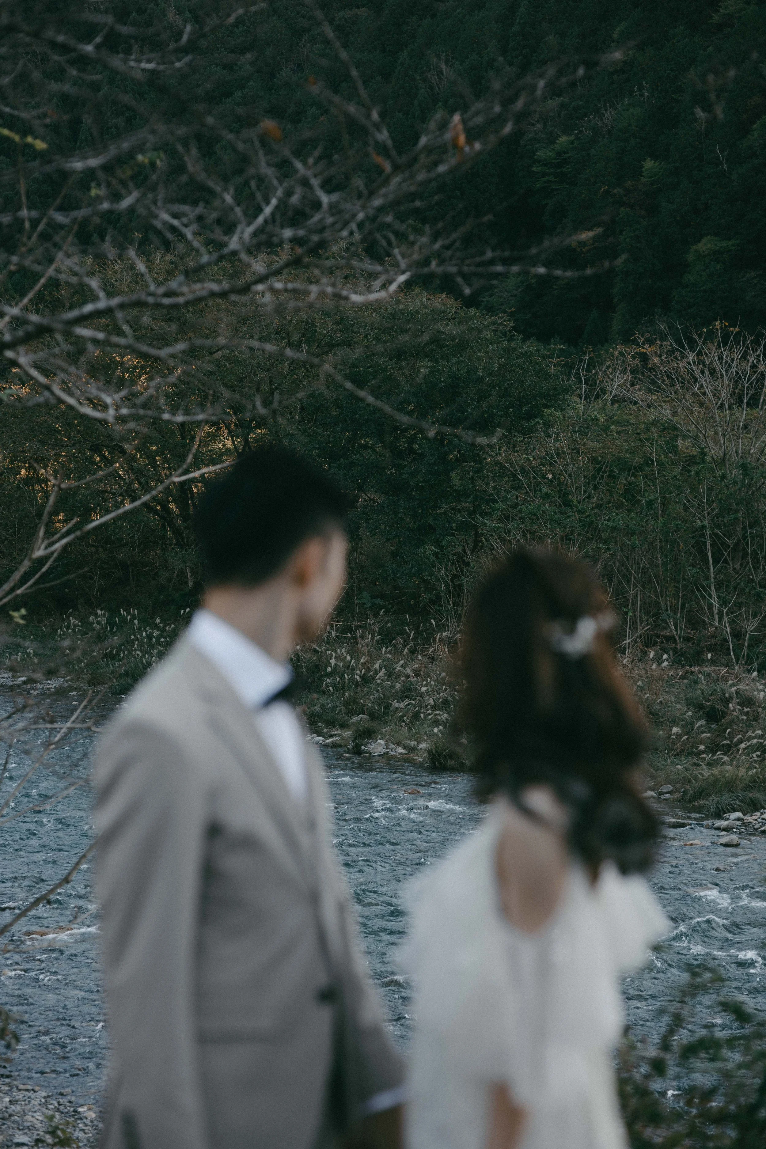 A couple dressed in wedding attire standing near a river with trees and mountains in the background.