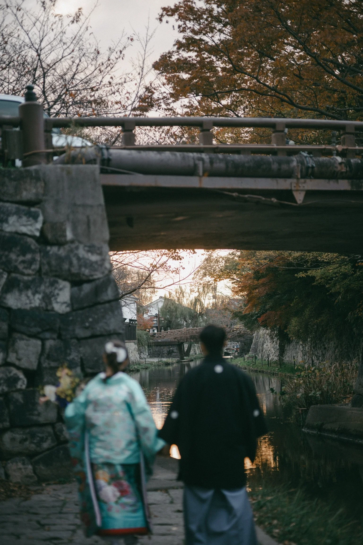 A couple in traditional Japanese clothing walking along a riverside under a bridge during autumn.