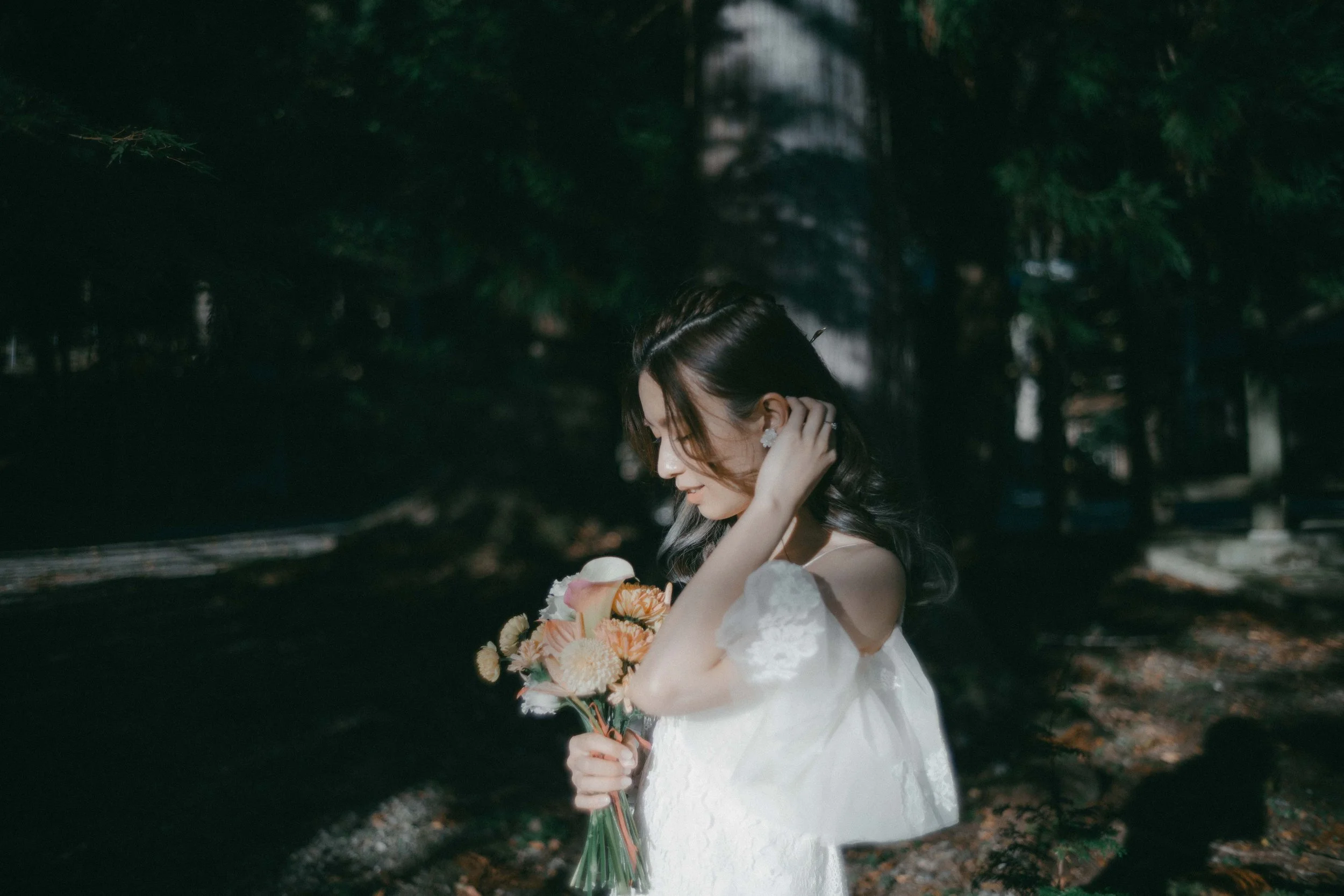 A woman in a white dress holding a bouquet of flowers in a forested area with sunlight filtering through trees.
