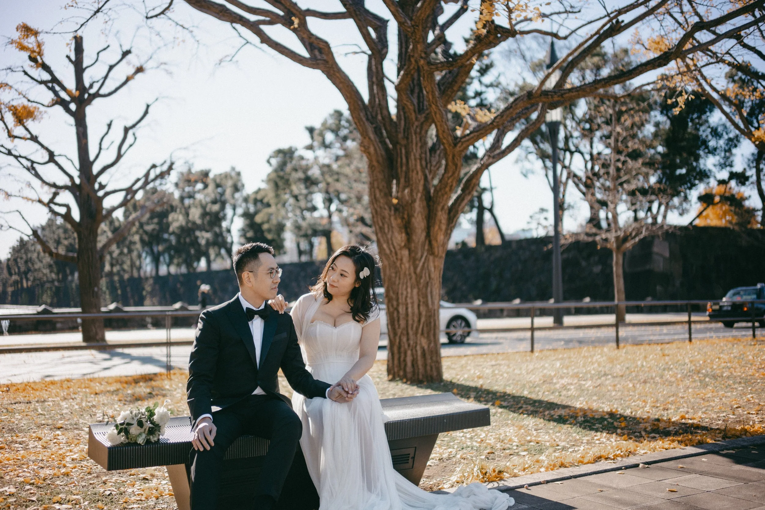 A bride and groom sitting on a park bench under a large tree, holding hands and gazing at each other. The bride wears a white wedding dress with hair accessories, and the groom wears a black tuxedo with glasses. There is a bouquet on the bench next t