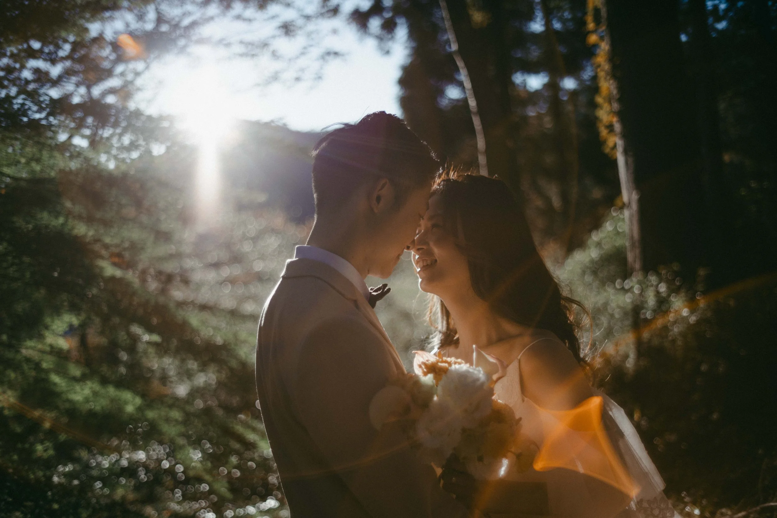 A couple in wedding attire holding a bouquet, standing close together with foreheads touching, in a forest with sunlight filtering through trees.