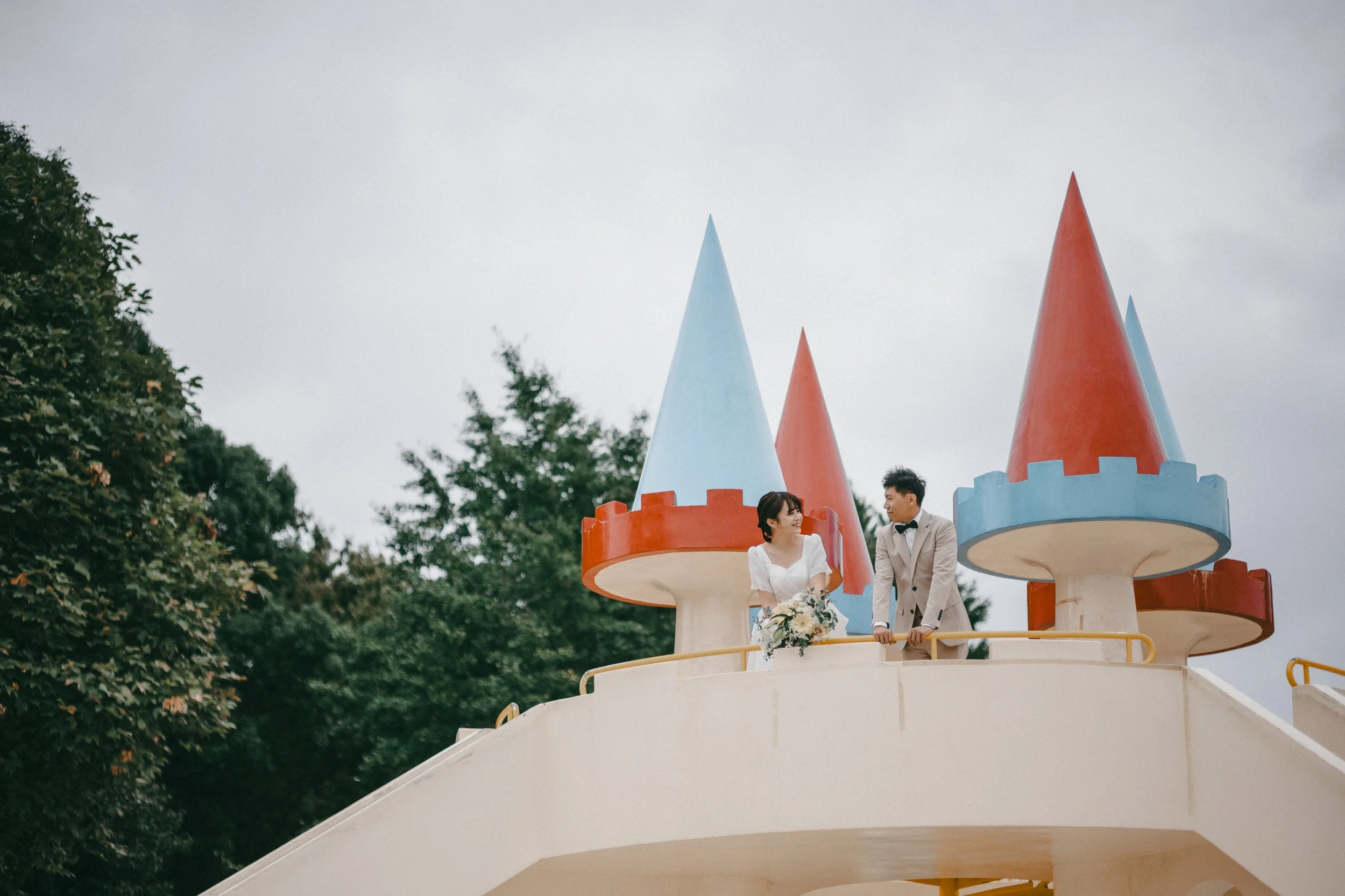 A bride and groom standing on a castle-themed platform outdoors, with the bride holding a bouquet of flowers and looking at the groom, against a backdrop of trees and an overcast sky.