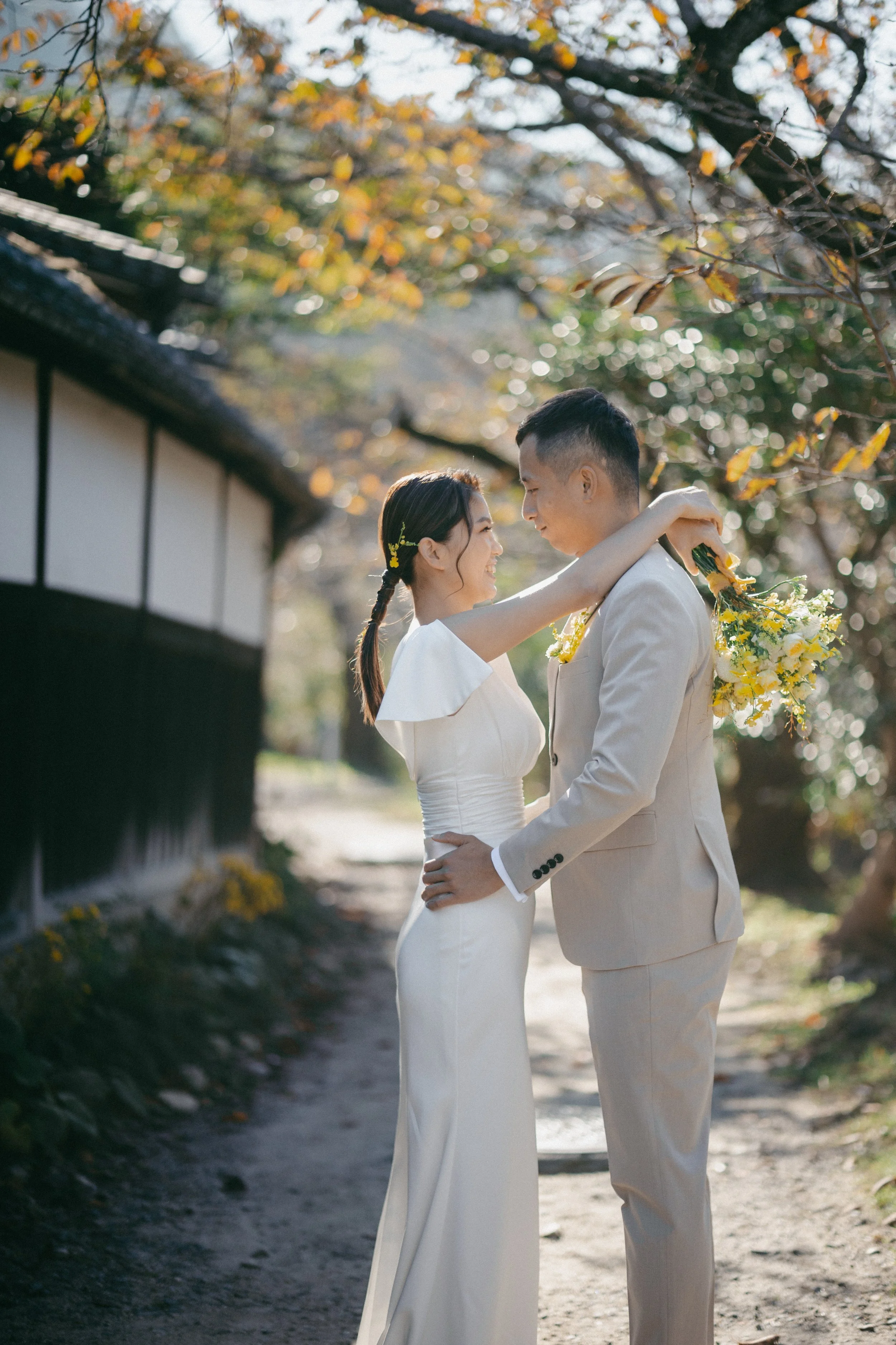 A bride and groom embrace outdoors, smiling at each other during their wedding, with trees and a traditional building in the background.
