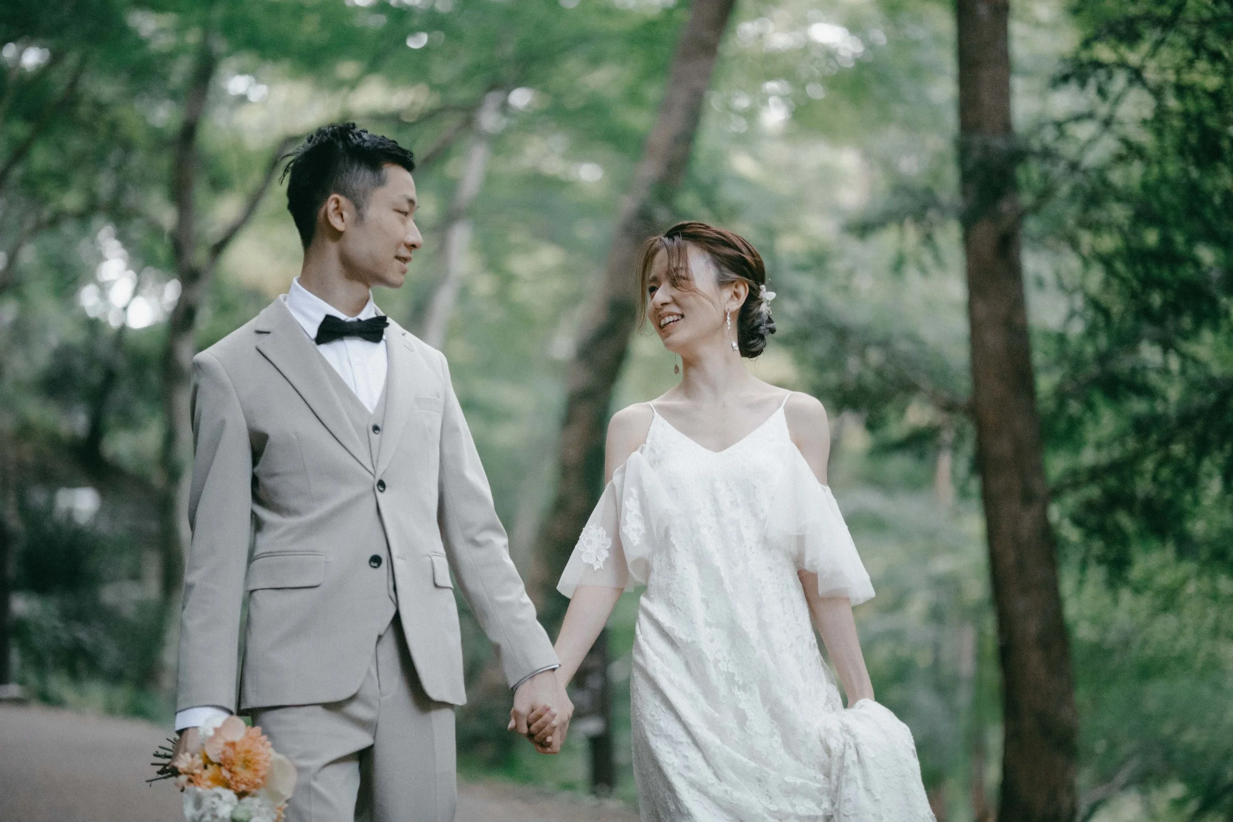 A couple walking in a forest, holding hands. The man is wearing a beige suit with a black bow tie and holding a bouquet. The woman is in a white lace wedding dress, smiling and looking at the man.