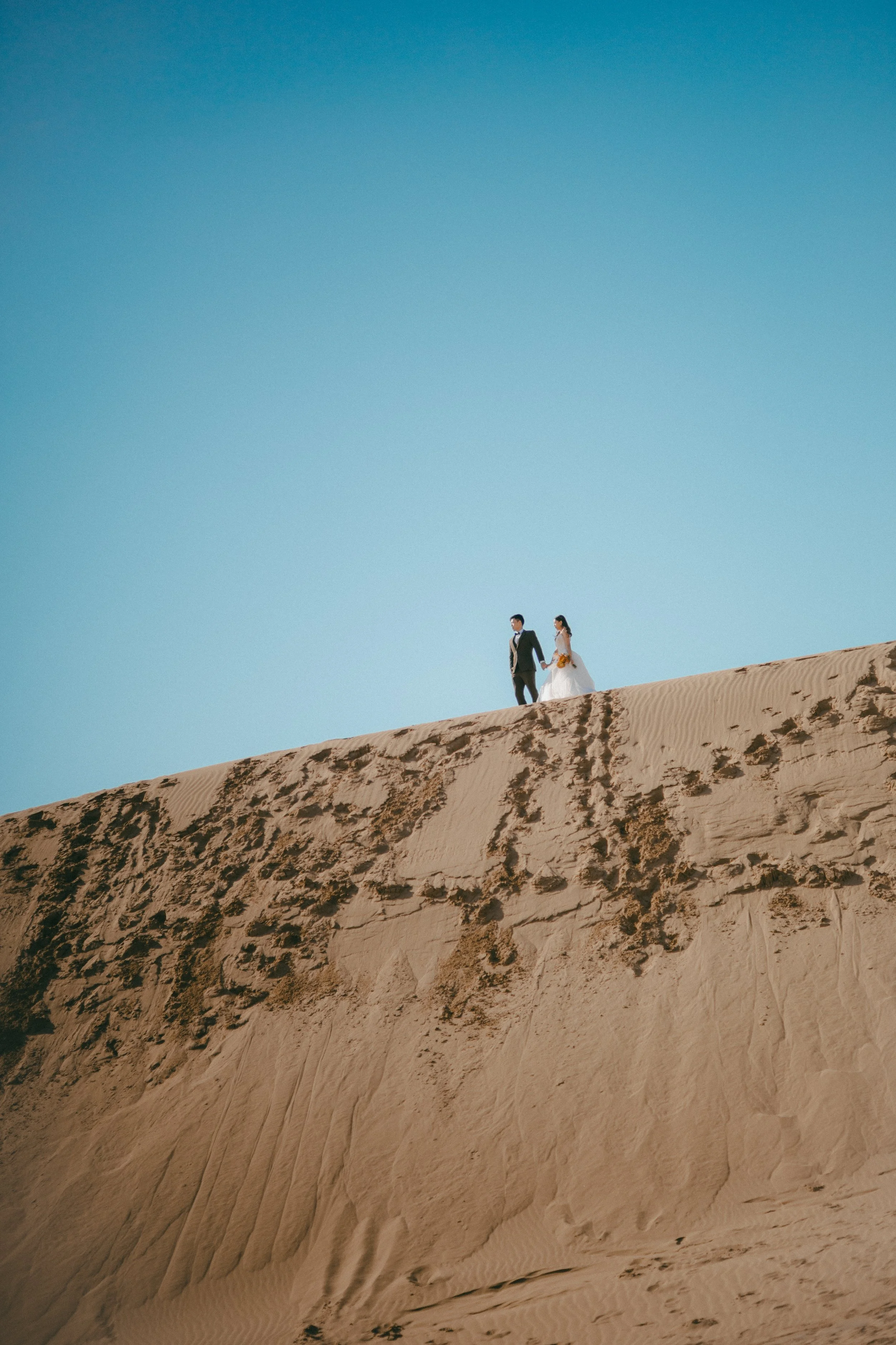 A bride and groom walking on top of a sand dune under a clear blue sky, dressed in wedding attire.