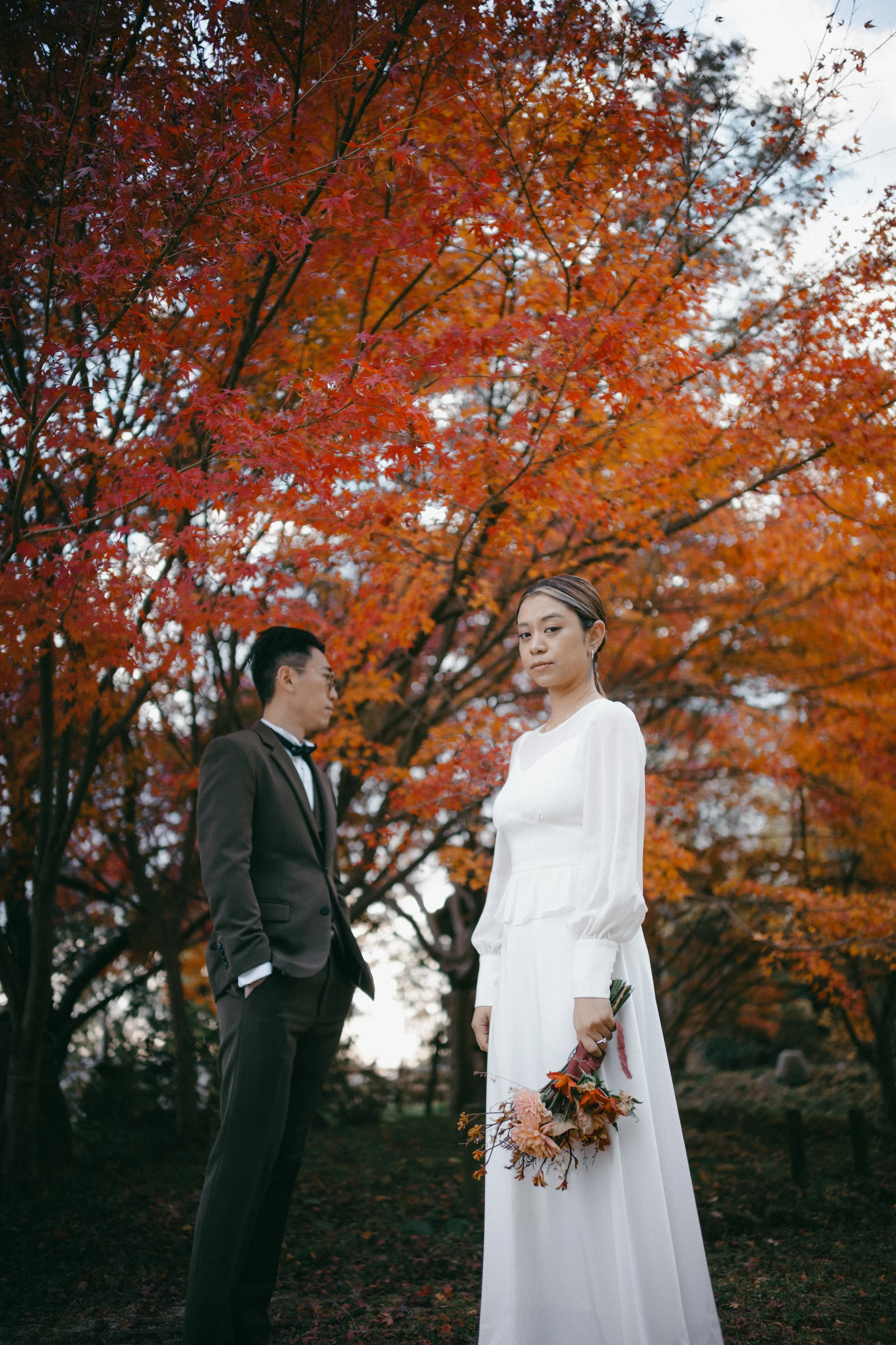 A woman in a white dress holding a bouquet of flowers, standing outdoors in front of a vibrant red-orange autumn tree, with a man in a dark suit and bowtie standing nearby and looking at her.