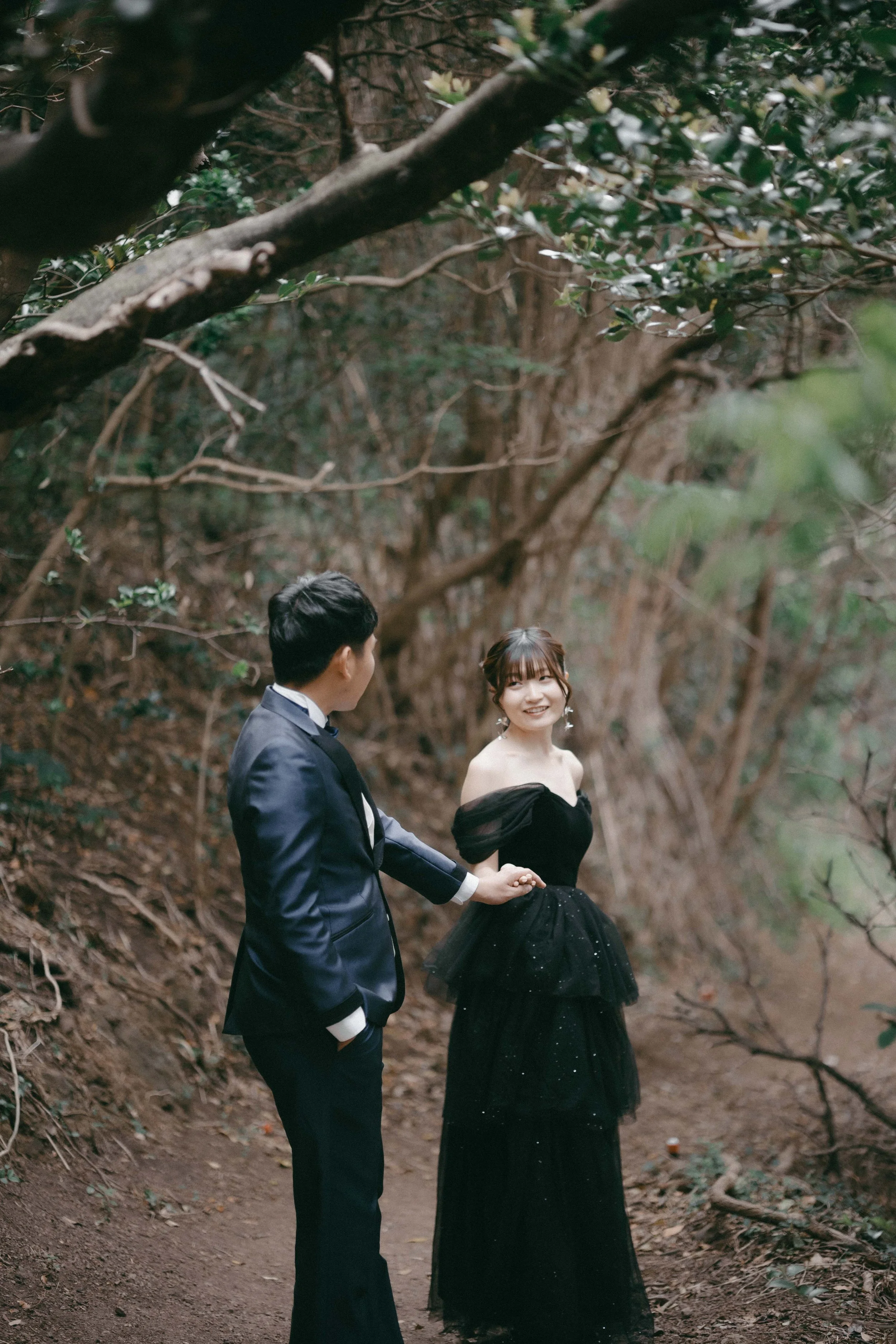 A couple in formal attire holding hands in a wooded area, with the woman smiling at the man.
