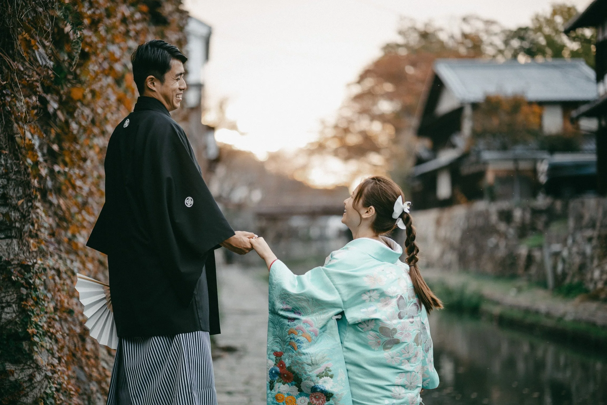 A couple in traditional Japanese attire exchanging a hand-held moment on a riverbank during autumn, with trees and houses in the background.