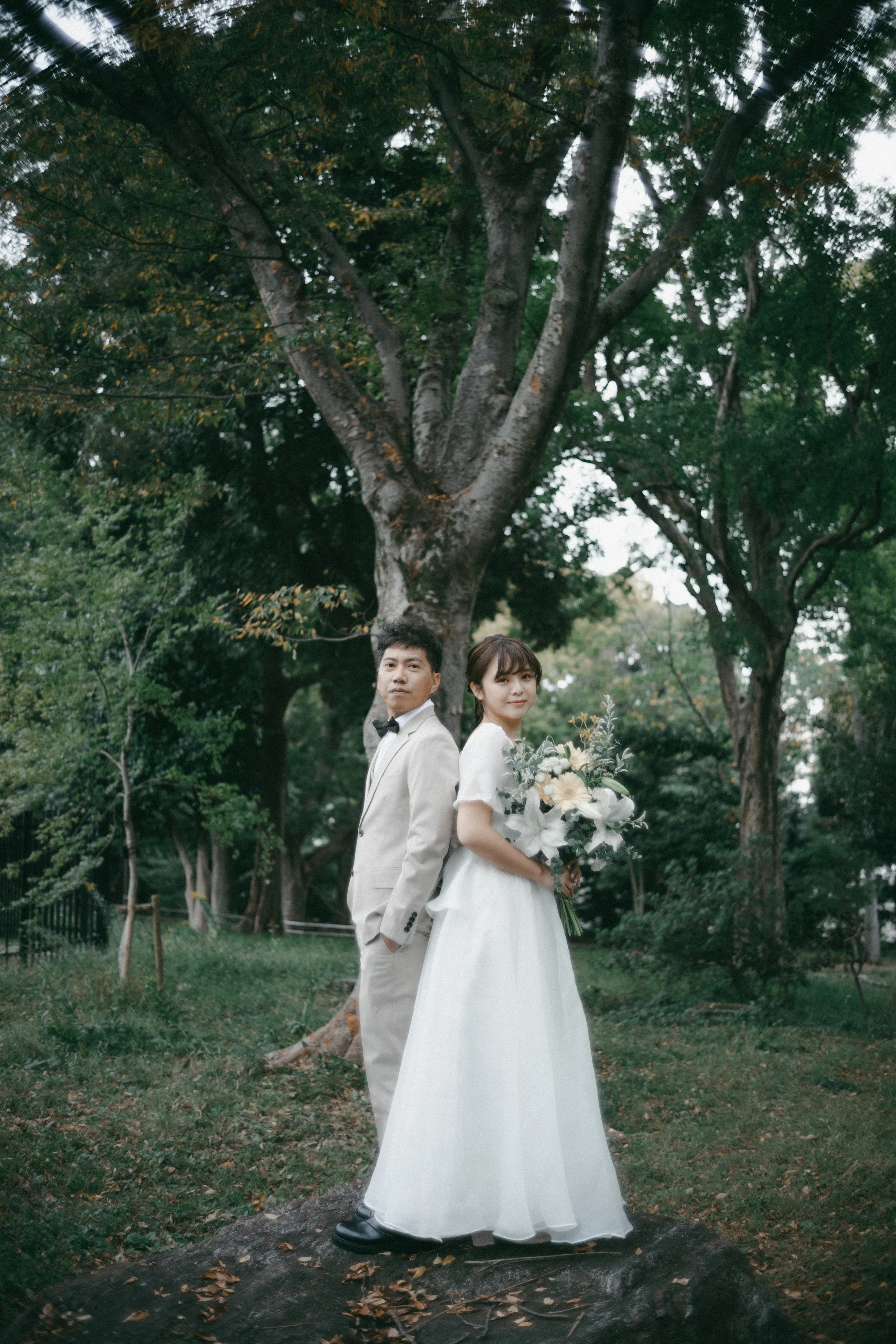 A bride and groom in wedding attire standing outdoors near a large tree and greenery, with the bride holding a bouquet of white flowers.