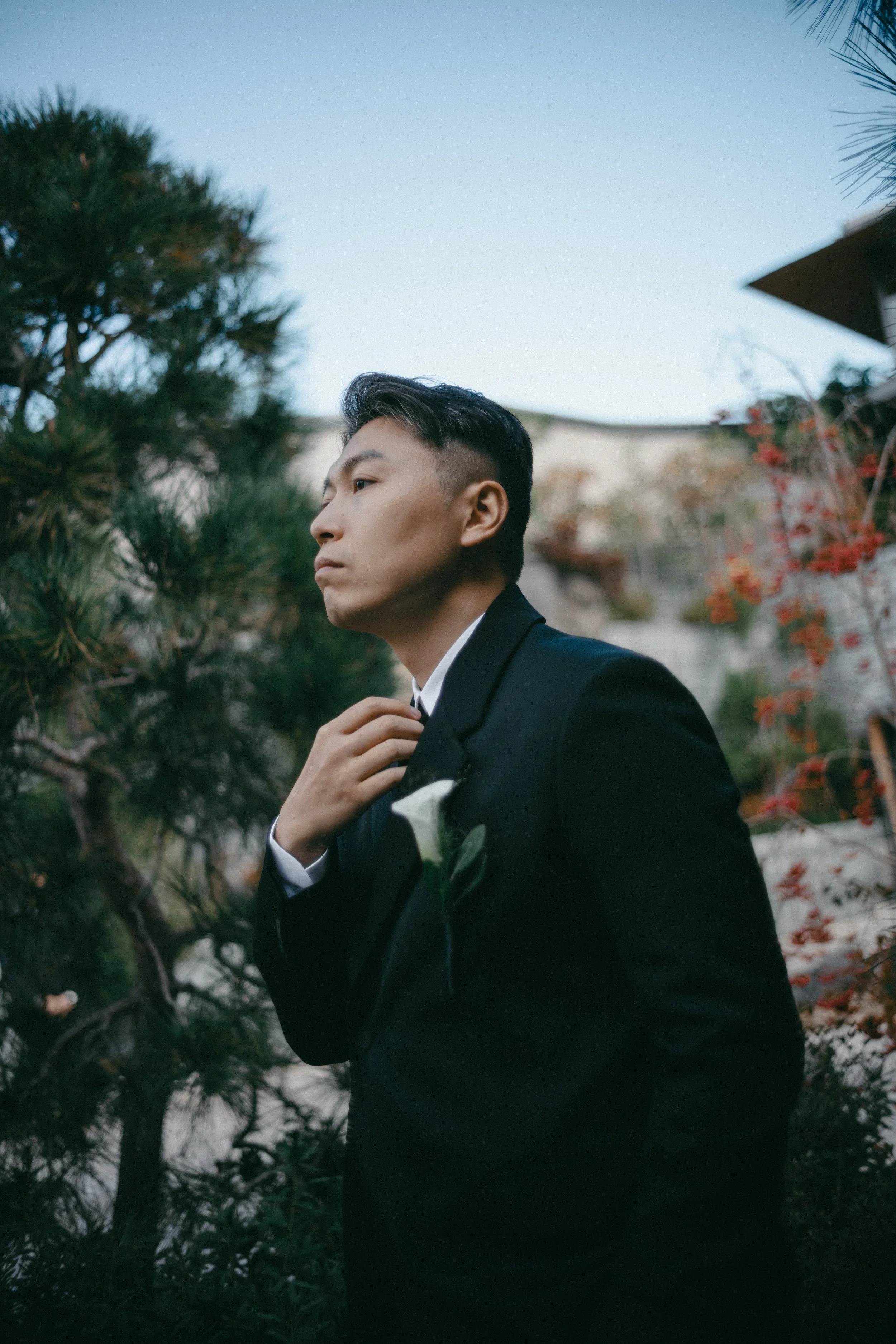 A man in a black suit with a white flower boutonniere, possibly a groom, standing outdoors among trees, adjusting his tie or collar, with a serious expression on his face.