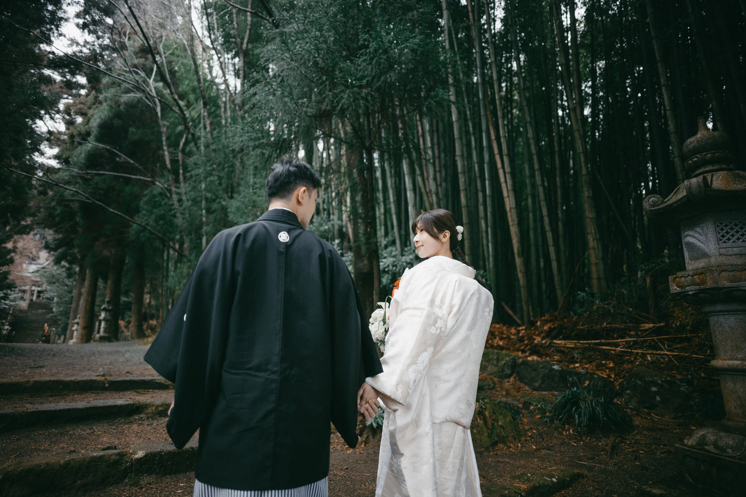A couple dressed in traditional Japanese wedding attire holding hands in a forested area with bamboo trees.
