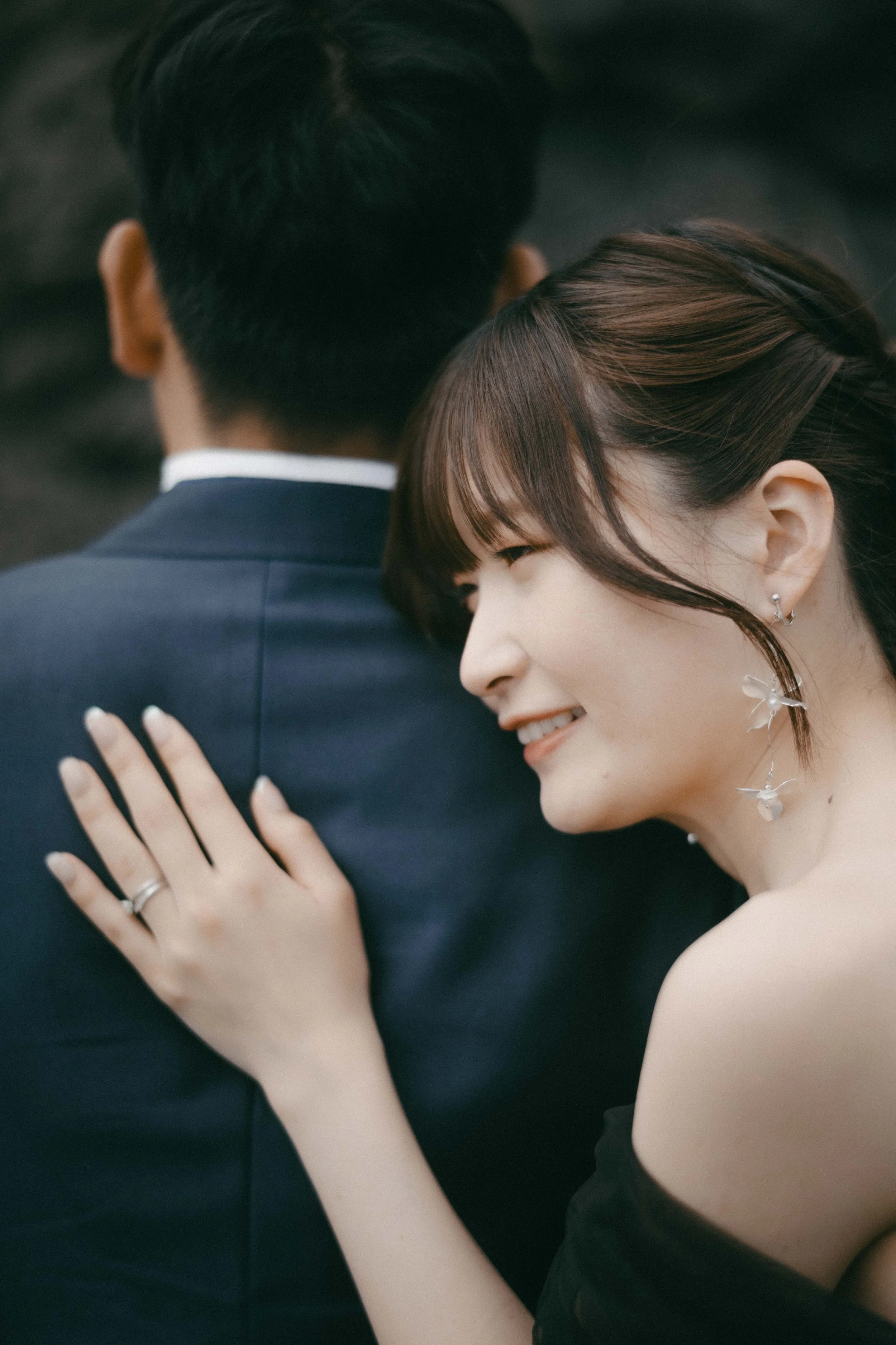 A woman with short hair and earrings is smiling and hugging a man in a suit from behind.