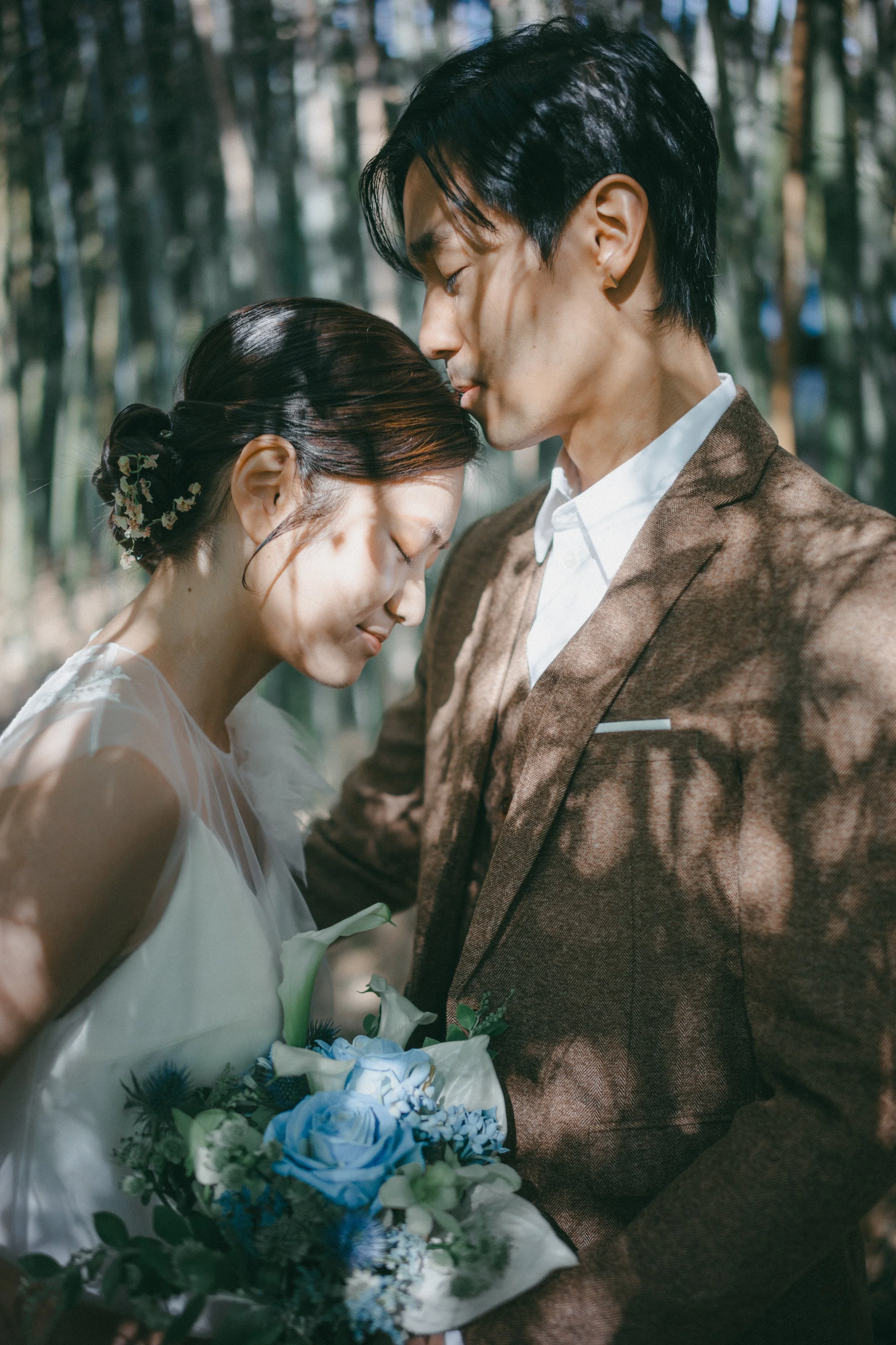 A bride and groom with their foreheads touching in a forest, the bride holding a bouquet of blue and white flowers, both with eyes closed and a peaceful expression.